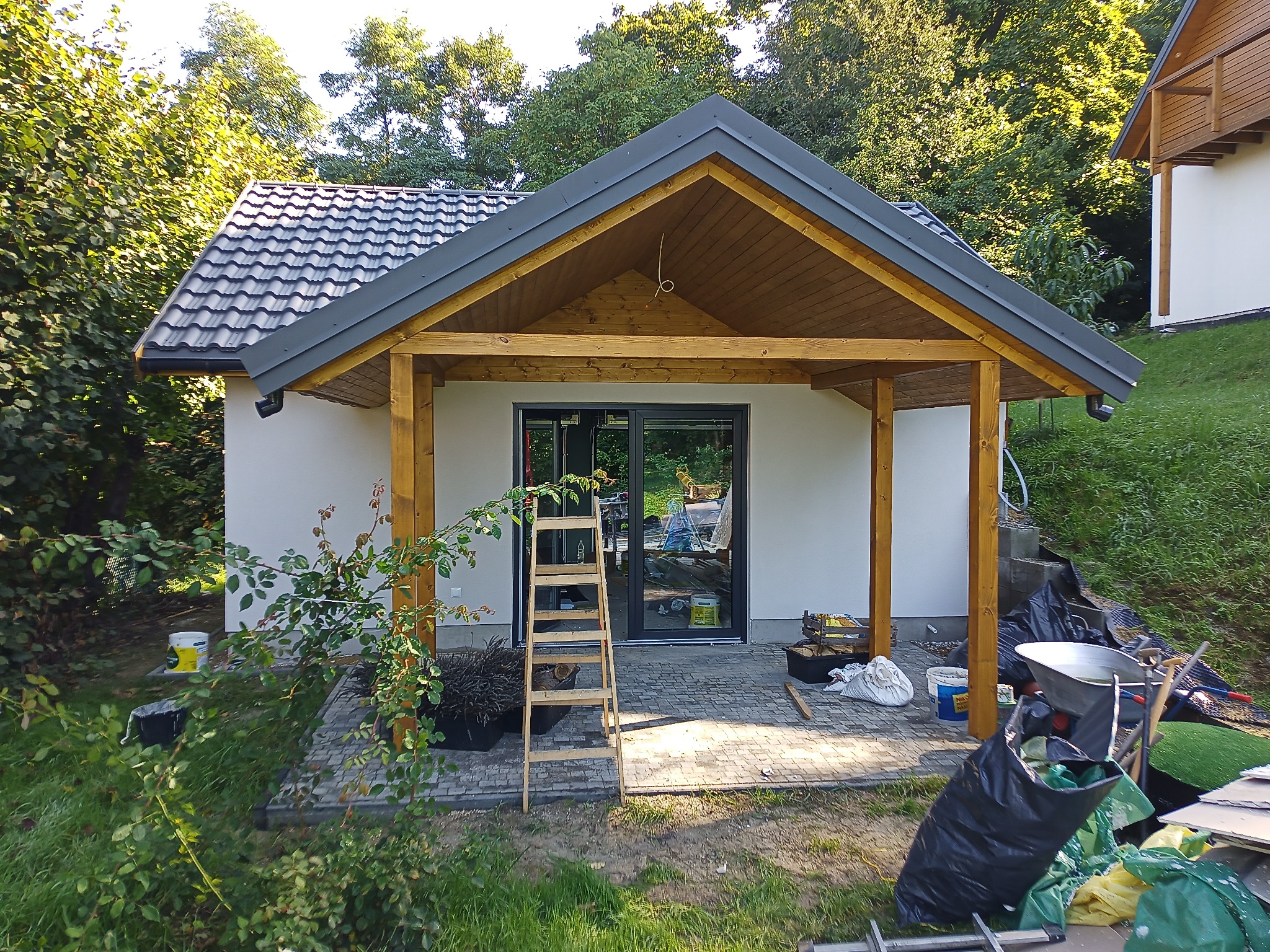 Niewielki budynek z drewnianą konstrukcją werandy, widoczne narzędzia i materiały budowlane, drabina oparta o przesuwne drzwi, rośliny w donicach z przodu.