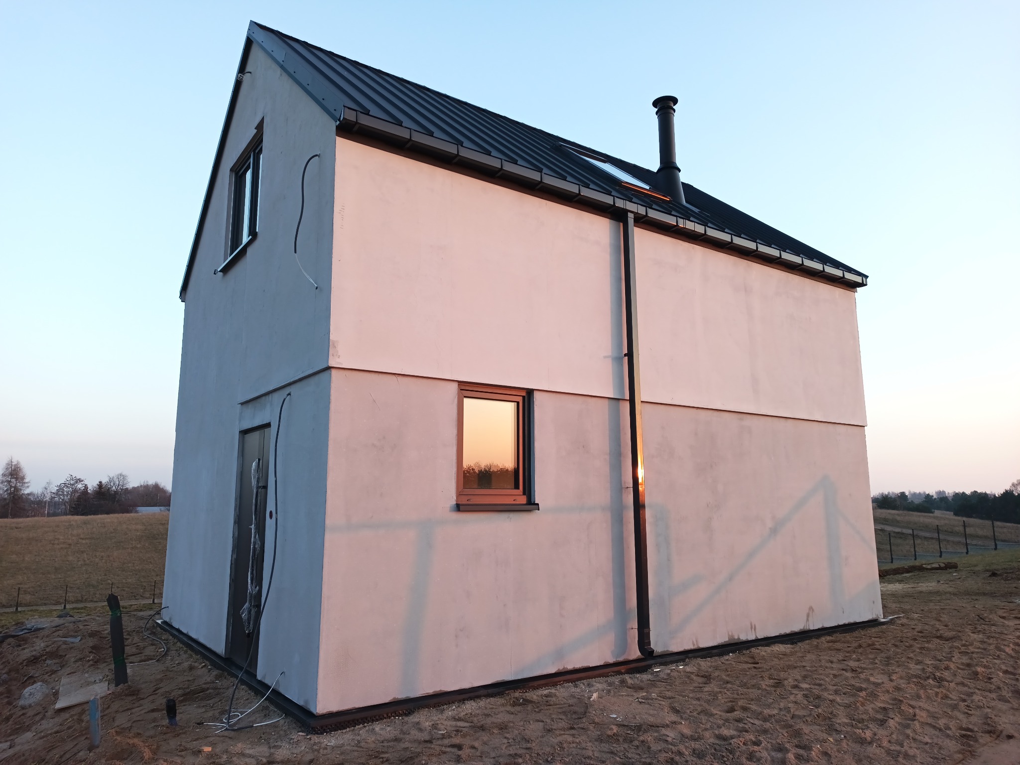 Nowoczesny dom o prostej bryle z płaskim dachem i czarną blachą, widoczny podczas zachodu słońca, z terenem wokół w trakcie zagospodarowywania.