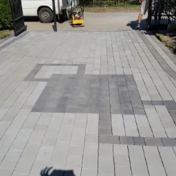 Ułożona kostka brukowa w odcieniach szarości z dekoracyjnym wzorem geometrycznym na podjeździe, z widocznym zagęszczarką i stosem kostki w tle.
