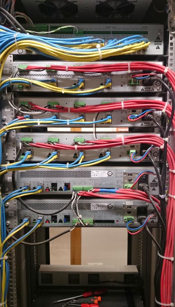 Szafa rack z zainstalowanymi modułami elektronicznymi Bosch, okablowanie w kolorach niebieskim, żółtym i czerwonym, widoczne narzędzia na półce poniżej.