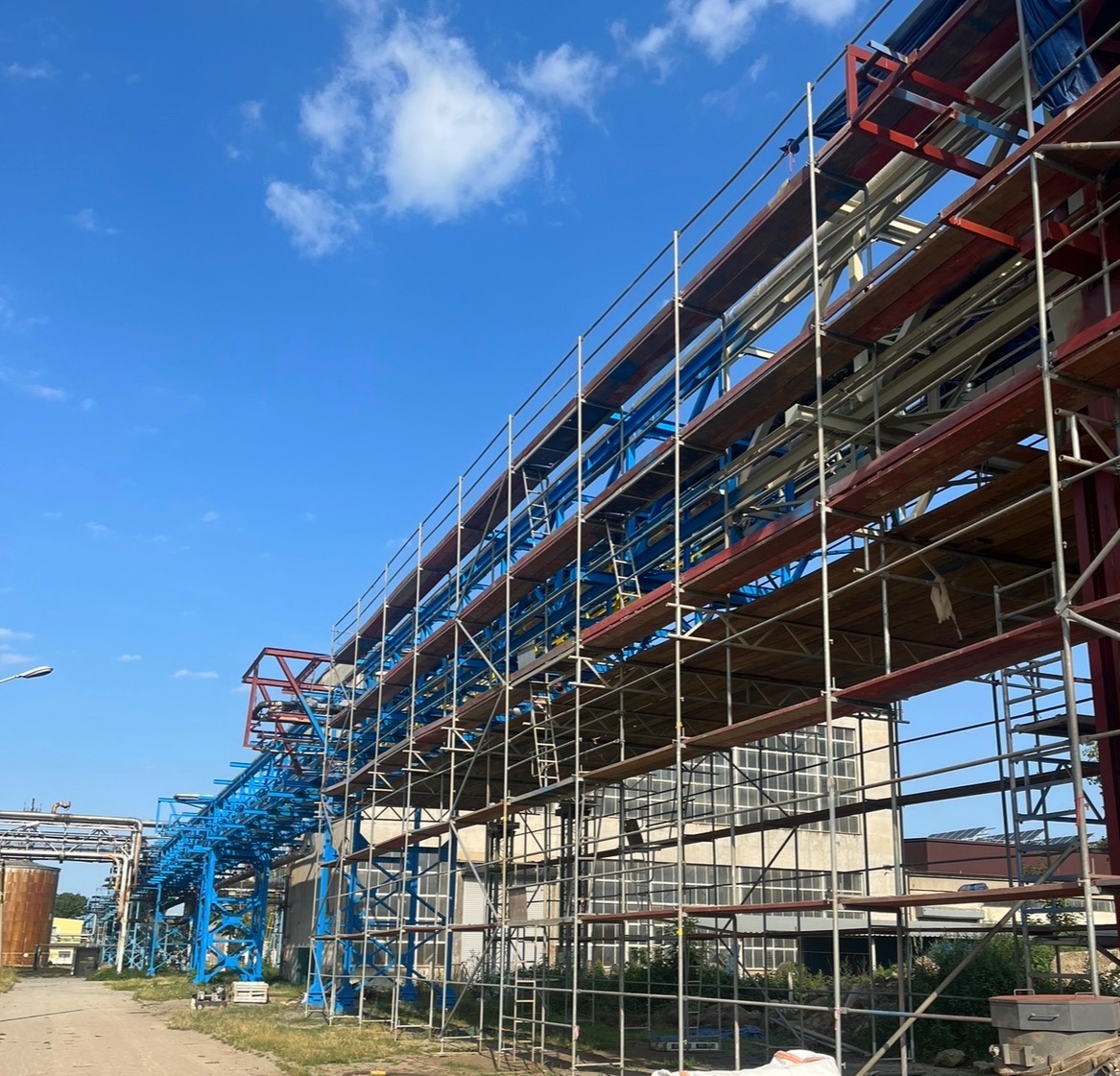 Konstrukcja stalowa z rusztowaniem, rury w kolorze niebieskim i czerwonym na tle błękitnego nieba. Widoczne elementy budynku przemysłowego.