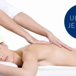 Kobieta z kwiatem we włosach relaksuje się podczas masażu pleców w jasnym otoczeniu, logo Ultra Jerzyka w prawym górnym rogu.