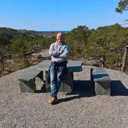 Mężczyzna oparty o kamienny stół piknikowy na wzgórzu z widokiem na las i morze w oddali.