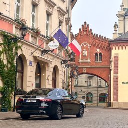 NEED NOW ADRIAN JASKÓLSKI - Czarny Mercedes parkuje na brukowanej ulicy przed Hotelem Polski w Krakowie, z flagami Polski i Unii Europejskiej powiewającymi nad wejściem i ozdobną, ceglaną bramą w tle.