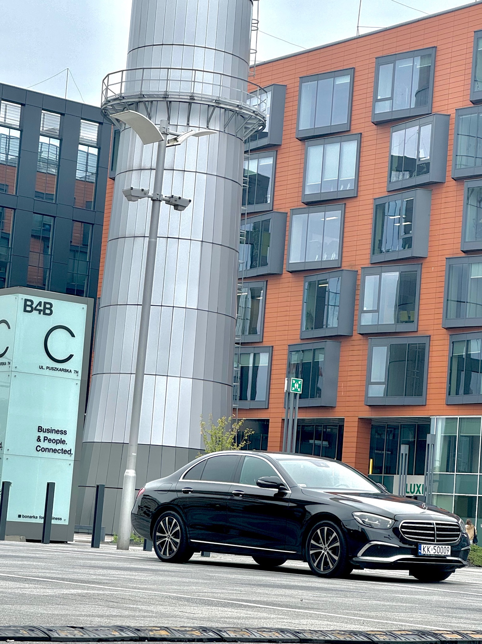 Czarny Mercedes E-Klasa na tle nowoczesnego biurowca z pomarańczową fasadą i srebrną konstrukcją przypominającą silos, tablica informacyjna B4B widoczna po lewej.