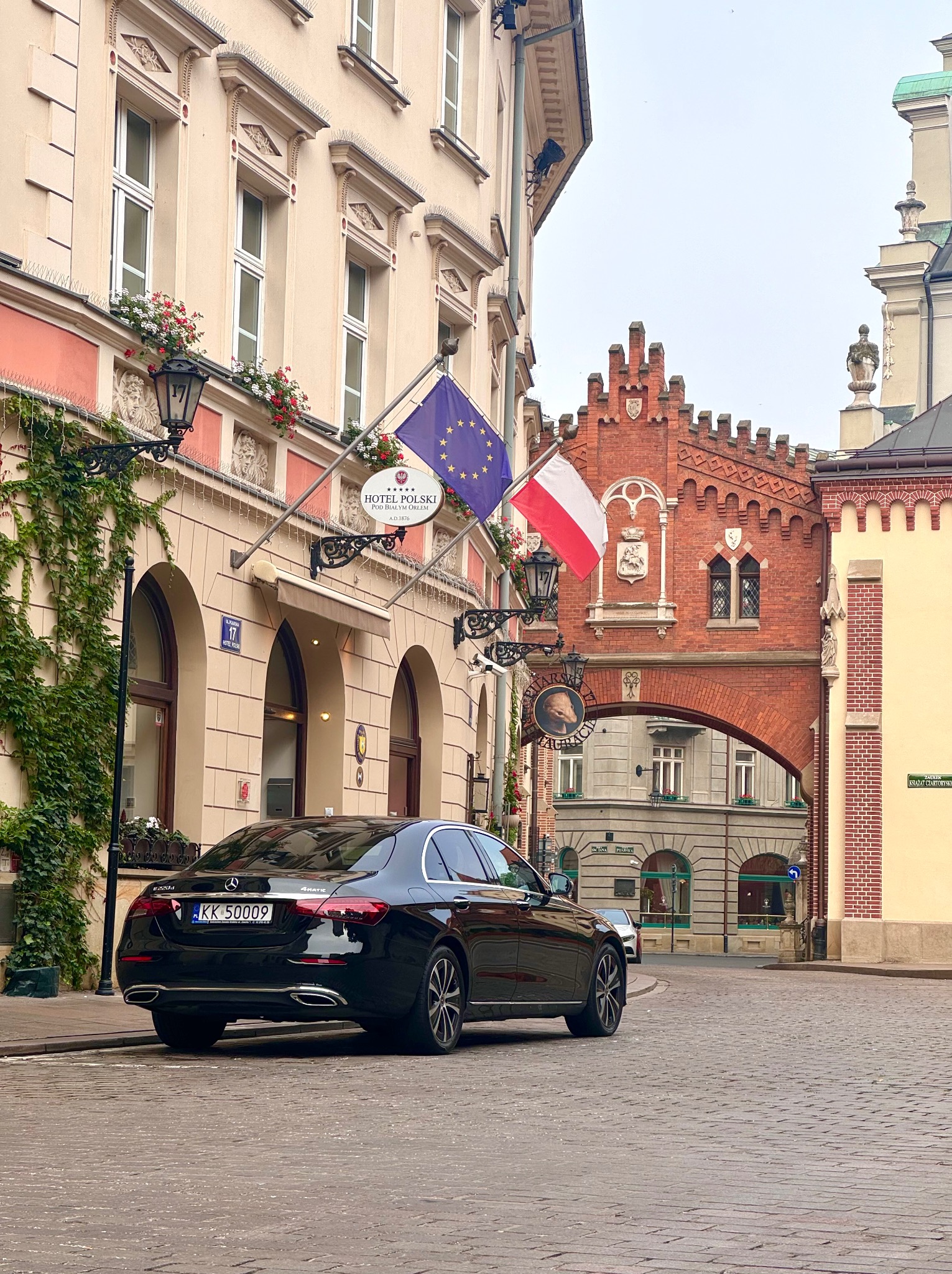 Czarny Mercedes parkuje na brukowanej ulicy przed Hotelem Polski w Krakowie, z flagami Polski i Unii Europejskiej powiewającymi nad wejściem i ozdobną, ceglaną bramą w tle.