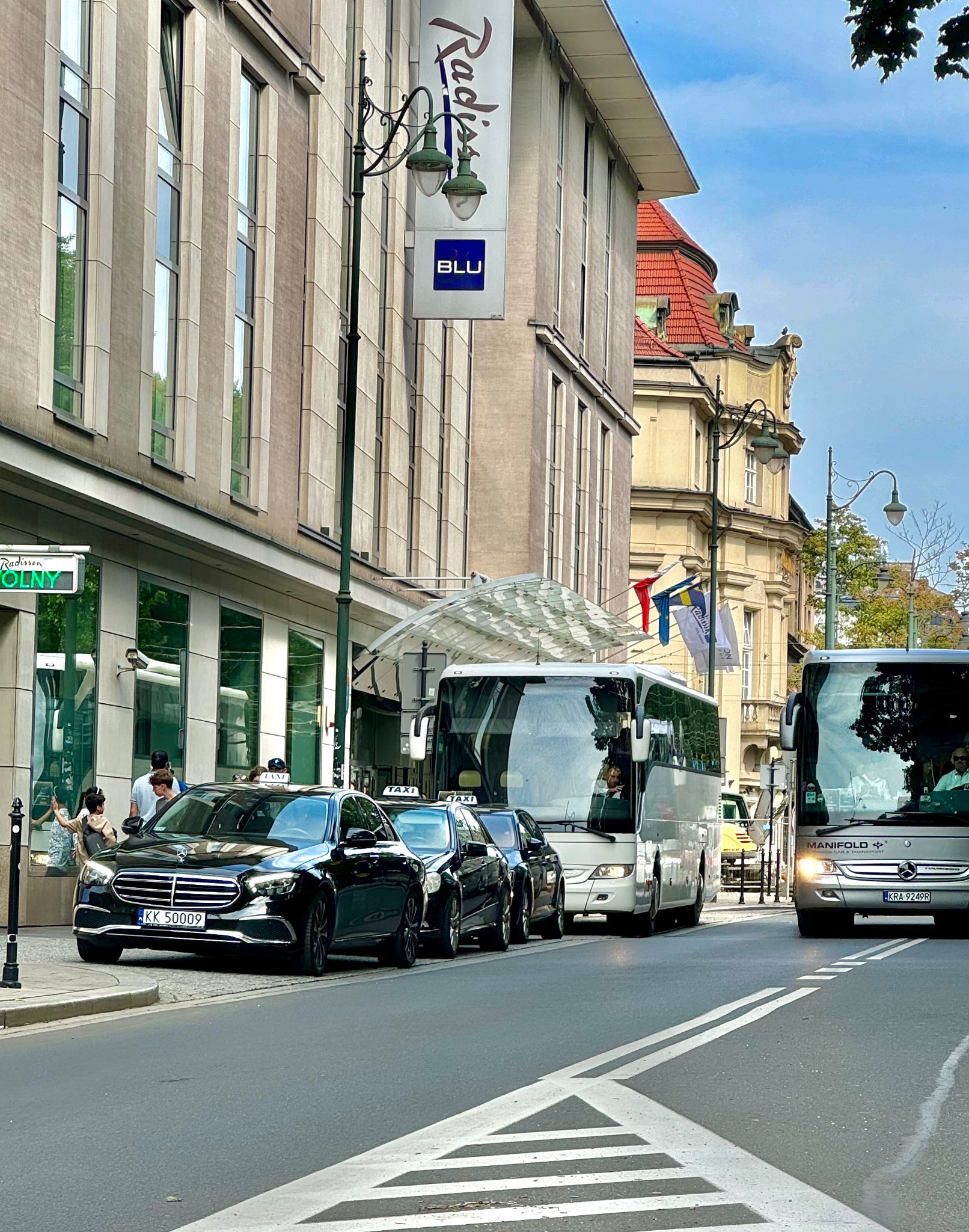 Czarne taksówki Mercedes i białe autokary stojące przed budynkiem Radisson Blu, widok z poziomu ulicy.