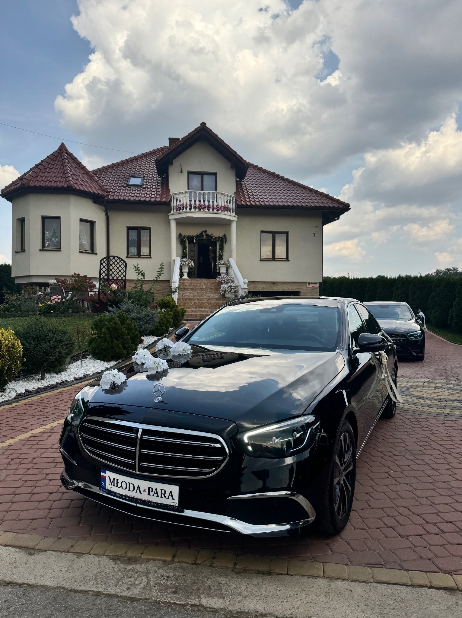 Czarny Mercedes udekorowany białymi kwiatami i tablicą 'Młoda Para' zaparkowany przed domem z balkonem przystrojonym kwiatami, w tle drugie auto.