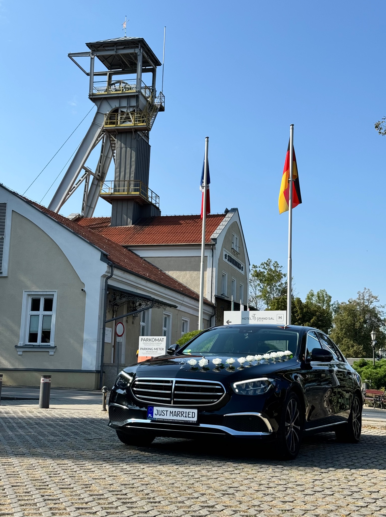 Czarny Mercedes udekorowany białymi różami na masce, z tablicą rejestracyjną 'JUST MARRIED', na tle szybu kopalnianego, flagi polskiej i niemieckiej oraz budynku hotelu Grand Sal.