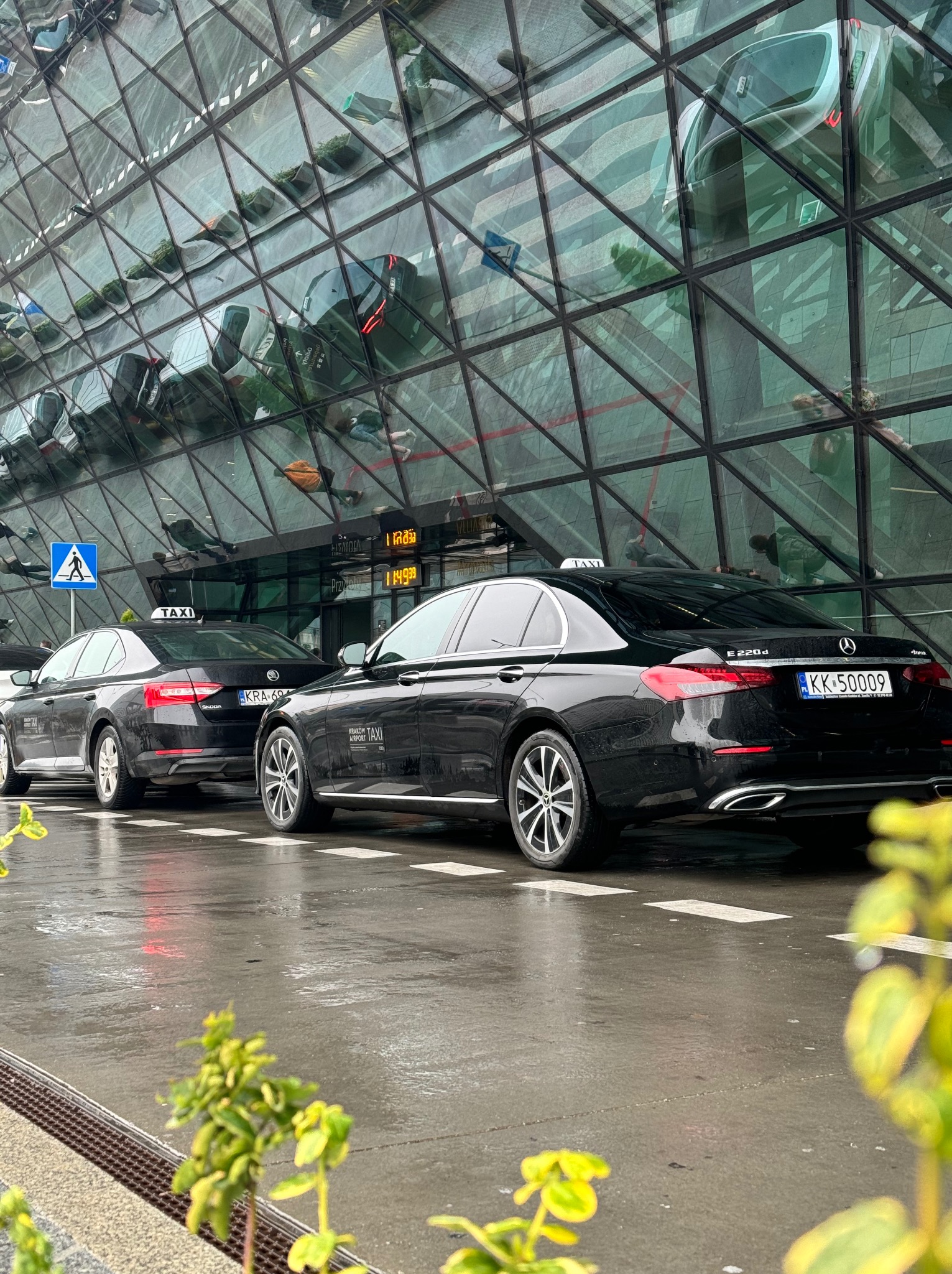 Dwa czarne samochody taxi, Mercedes i Skoda, stojące przed nowoczesnym budynkiem z refleksyjną szklaną fasadą. Na chodniku mokro po deszczu. Widoczny znak drogowy z symbolem pieszego.