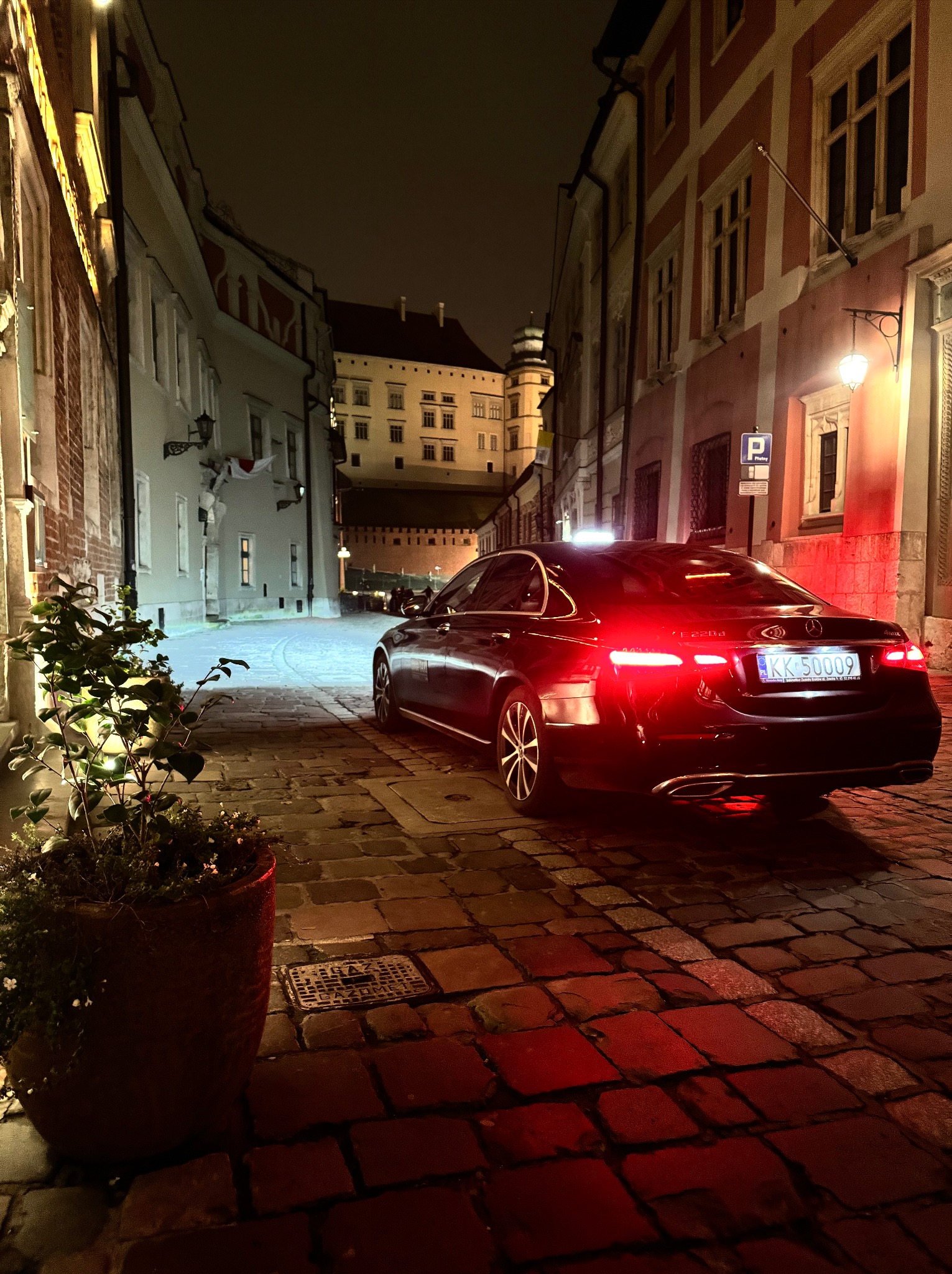 Czarny samochód osobowy Mercedes E220d z włączonymi światłami tylnymi, zaparkowany na brukowanej uliczce w Krakowie nocą, z widokiem na Wawel w tle.
