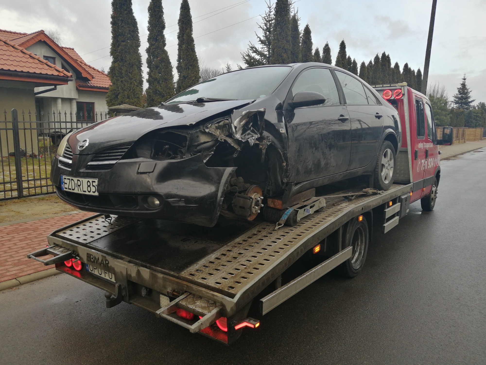 Czarny, uszkodzony samochód osobowy marki Nissan Primera na platformie czerwonej lawety z włączonymi światłami pozycyjnymi. Laweta zaparkowana na mokrej, asfaltowej drodze w pobliżu domów...