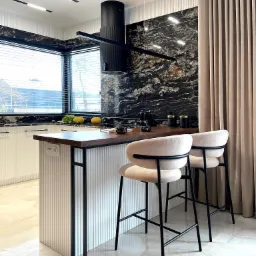 Nowoczesna kuchnia z wyspą, czarnym marmurem na ścianie, minimalistycznym okapem i beżowymi hokerami, oświetlona listwami LED.