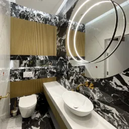 Nowoczesna łazienka z czarno-białymi marmurowymi płytkami, złotymi akcentami, wiszącą toaletą i umywalką nablatową na długim blacie, oświetlona okrągłym lustrem z LED.