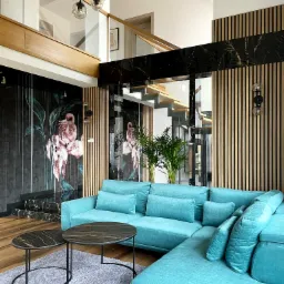 Turkusowy narożnik w salonie z akcentami czarnego marmuru i drewnianych lameli, ściana z motywem flamingów, nowoczesna lampa wisząca i schody z przeszkloną balustradą.