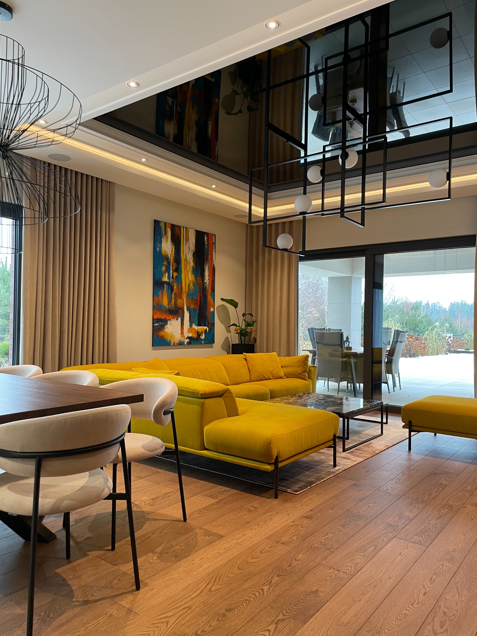 Nowoczesny salon z żółtą sofą modułową, abstrakcyjnym obrazem na ścianie i geometryczną lampą sufitową odbijającą się w czarnym lustrzanym suficie.