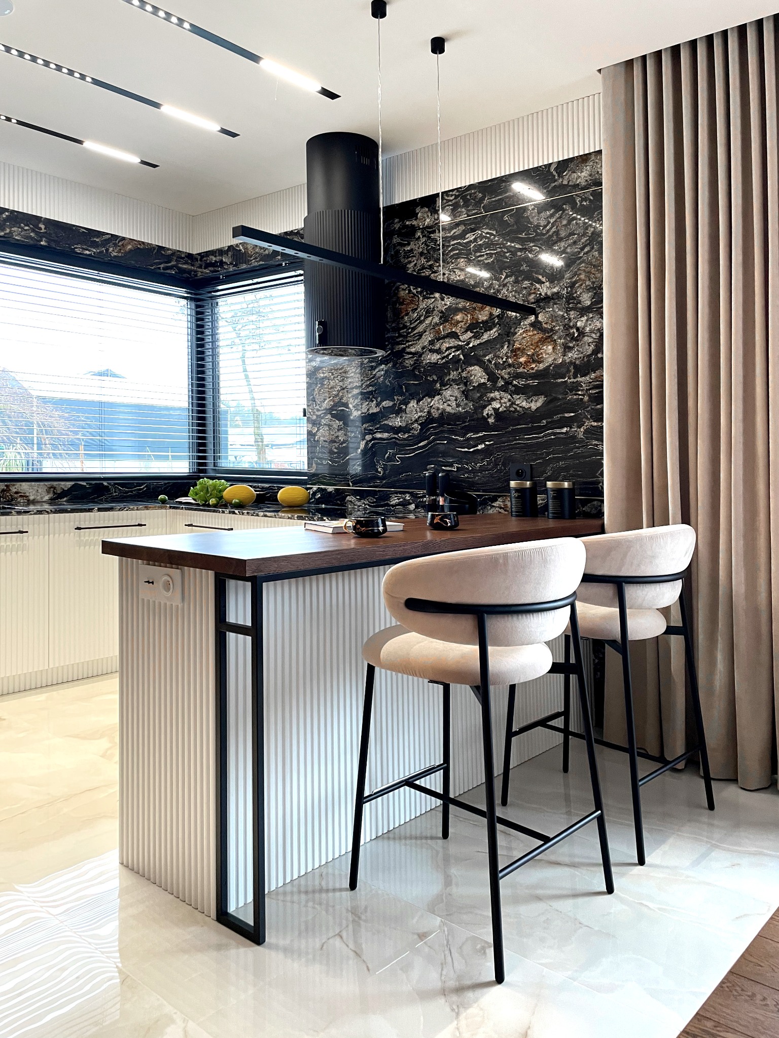 Nowoczesna kuchnia z wyspą, czarnym marmurem na ścianie, minimalistycznym okapem i beżowymi hokerami, oświetlona listwami LED.