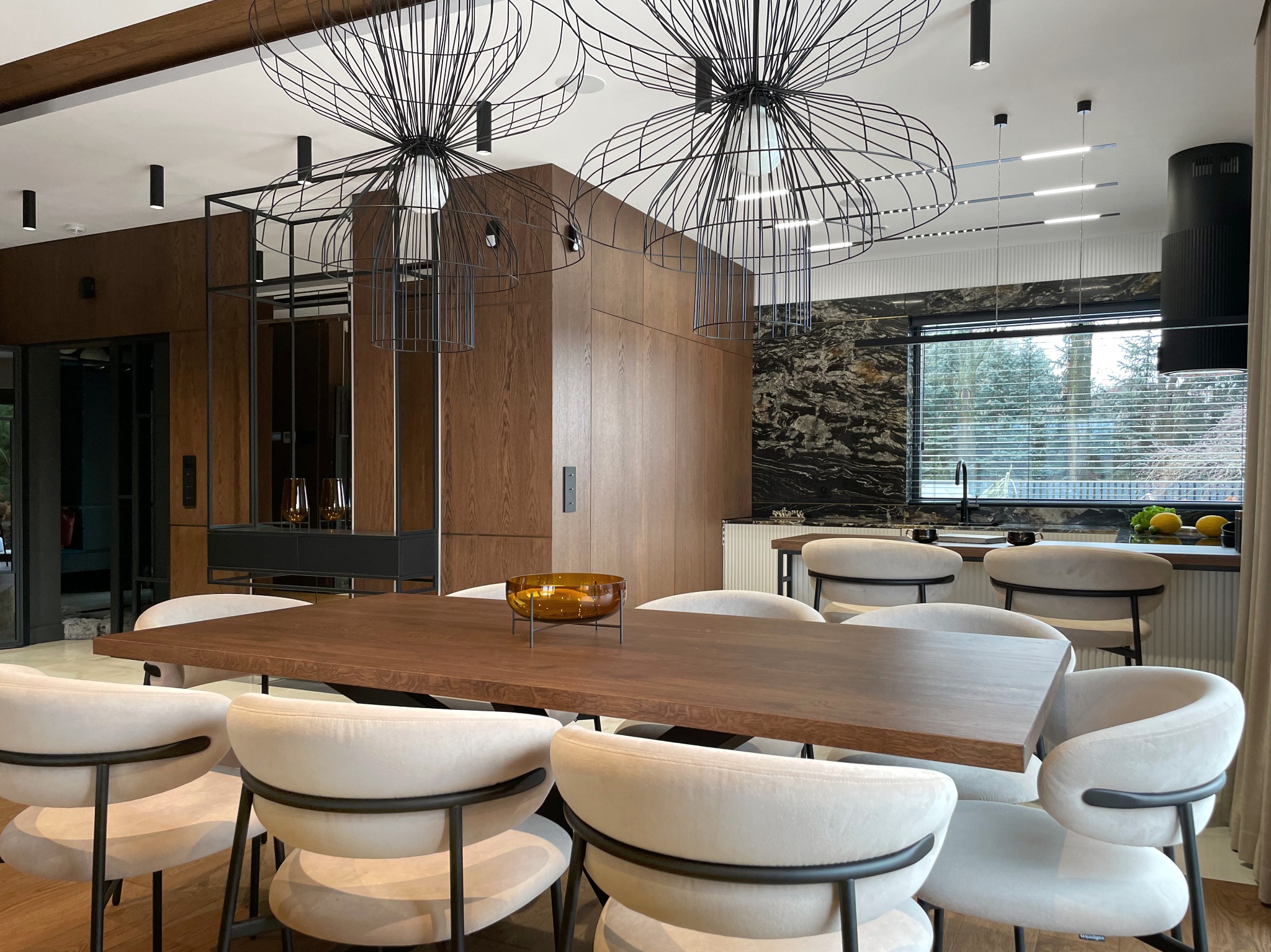 Nowoczesna jadalnia z drewnianym stołem otoczonym krzesłami tapicerowanymi w jasnym odcieniu, nad stołem wiszą ażurowe, metalowe lampy, w tle widoczna kuchnia z czarnym blatem i marmurową ścianą.