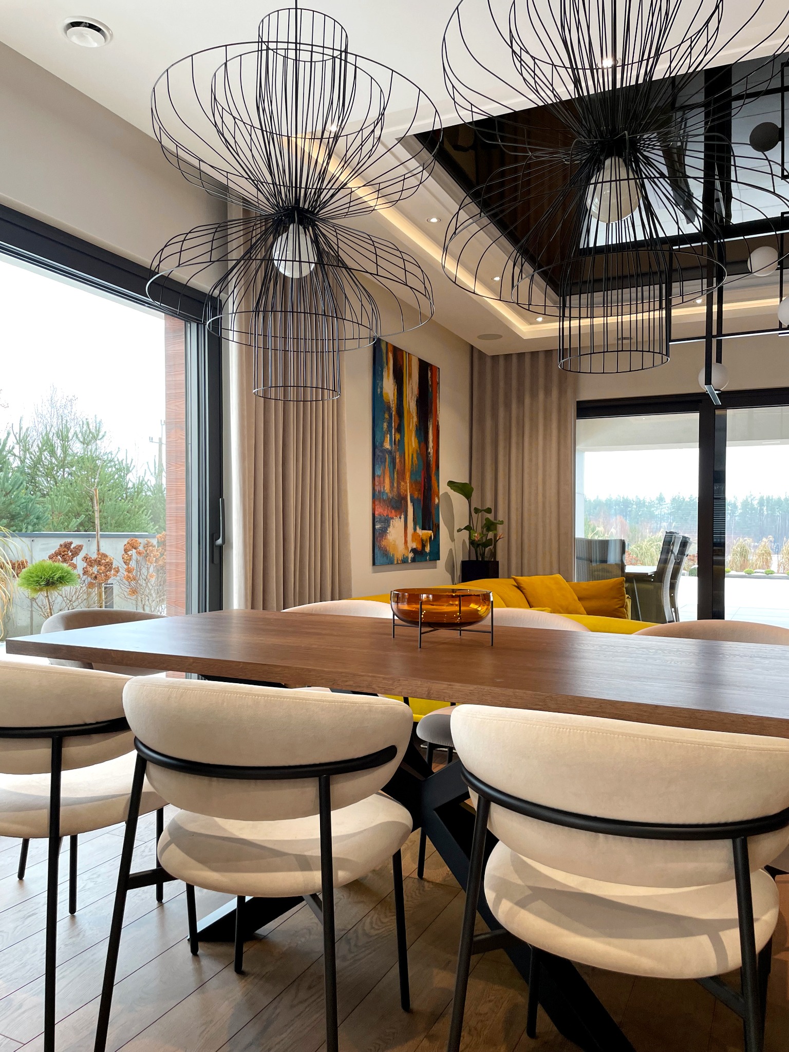 Nowoczesna jadalnia z dużym drewnianym stołem, krzesłami z jasną tapicerką i czarnymi nogami, wiszącymi lampami o ażurowej konstrukcji, abstrakcyjnym obrazem na ścianie i widokiem na ogród przez...