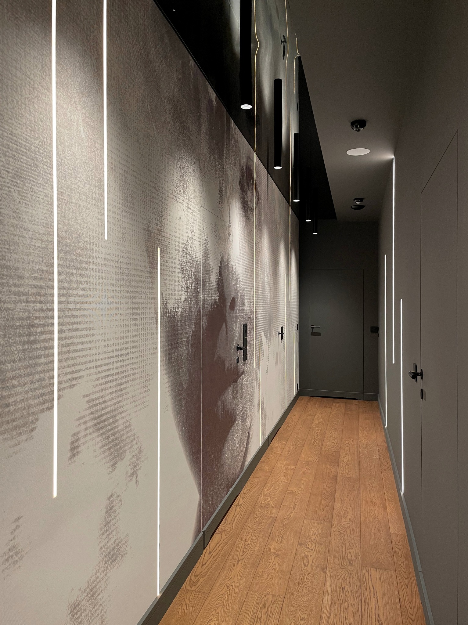 Nowoczesny korytarz z drewnianą podłogą, ścianą z abstrakcyjnym portretem podświetloną pionowymi paskami LED i czarnymi lampami sufitowymi.