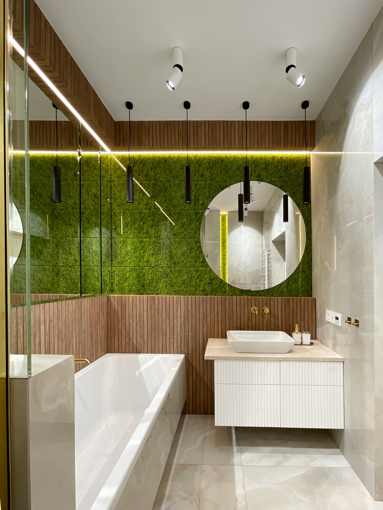 Nowoczesna łazienka z wanną, umywalką nablatową i ścianą z mchu, oświetlona wiszącymi lampami i punktowymi reflektorami.