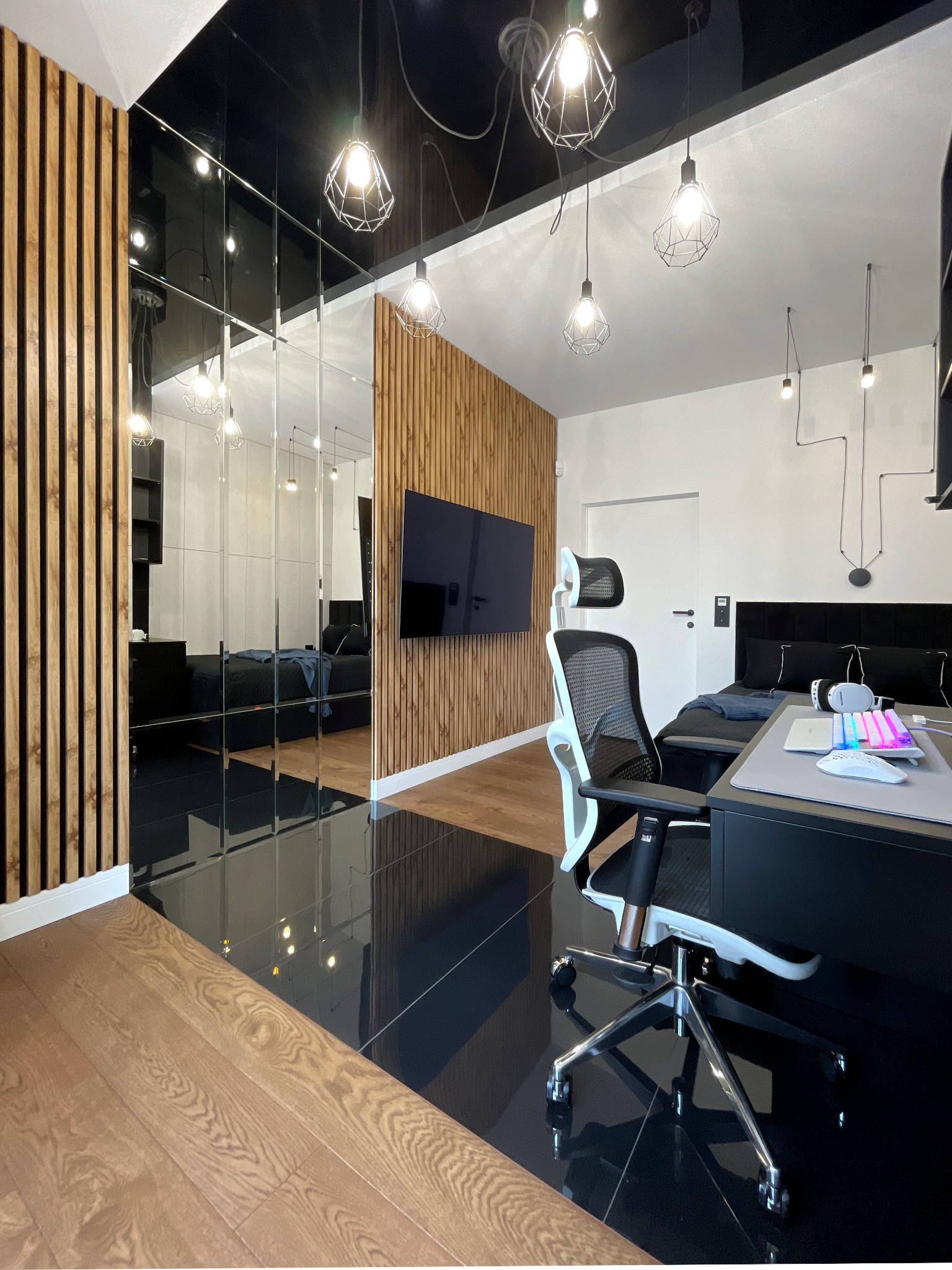 Nowoczesny pokój z czarną podłogą i drewnianymi panelami, biurko z komputerem i krzesłem, odbicie sypialni w lustrzanej ścianie, geometryczne lampy wiszące