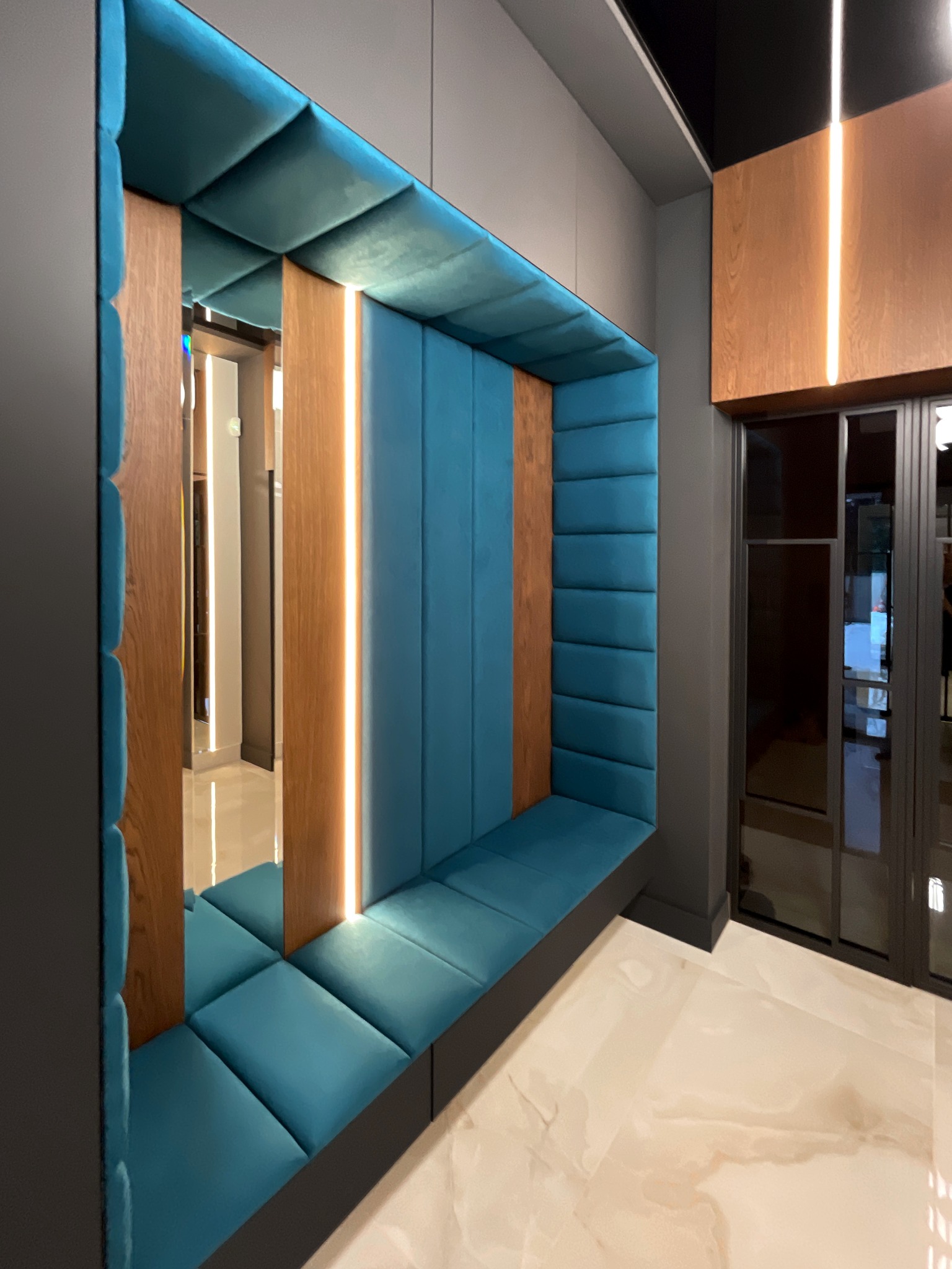 Wnęka w ścianie z tapicerowanym siedziskiem w kolorze turkusowym, oświetlona pionowymi pasami LED, z drewnianymi elementami dekoracyjnymi, widok na korytarz z lustrzanym odbiciem i przeszklone drzwi.