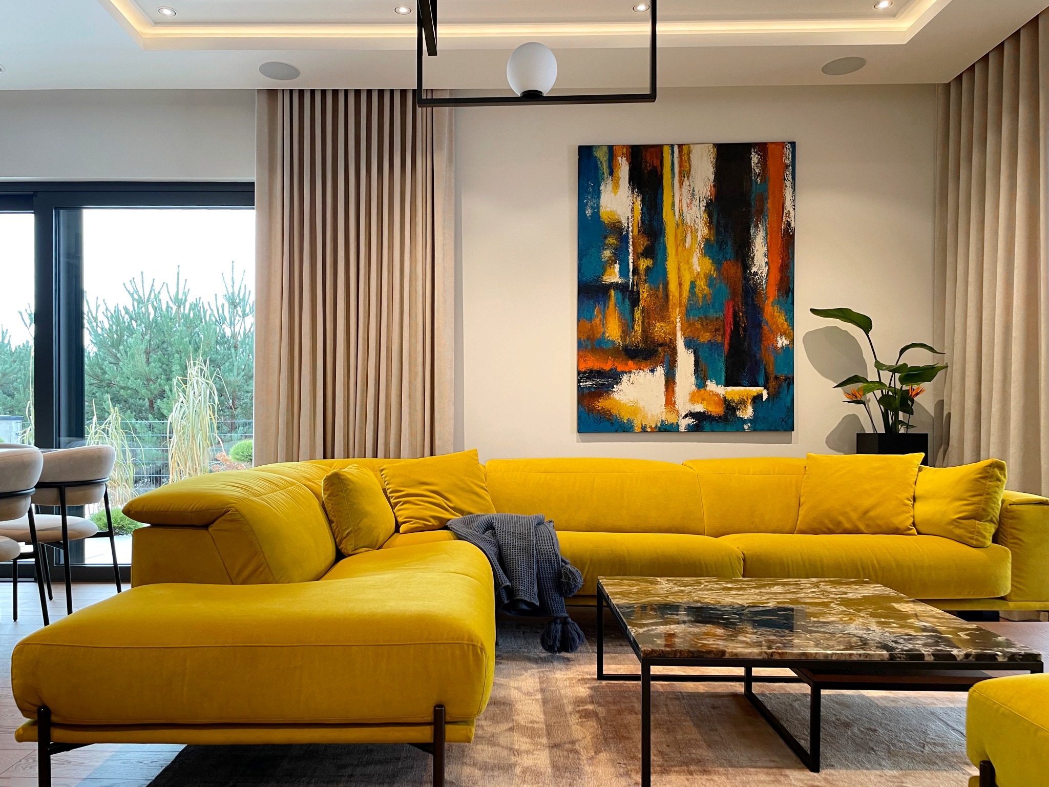 Salon z narożnikiem w odcieniu musztardowej żółci, abstrakcyjny obraz na ścianie, stolik kawowy z marmurowym blatem i widok na zieleń za oknem.