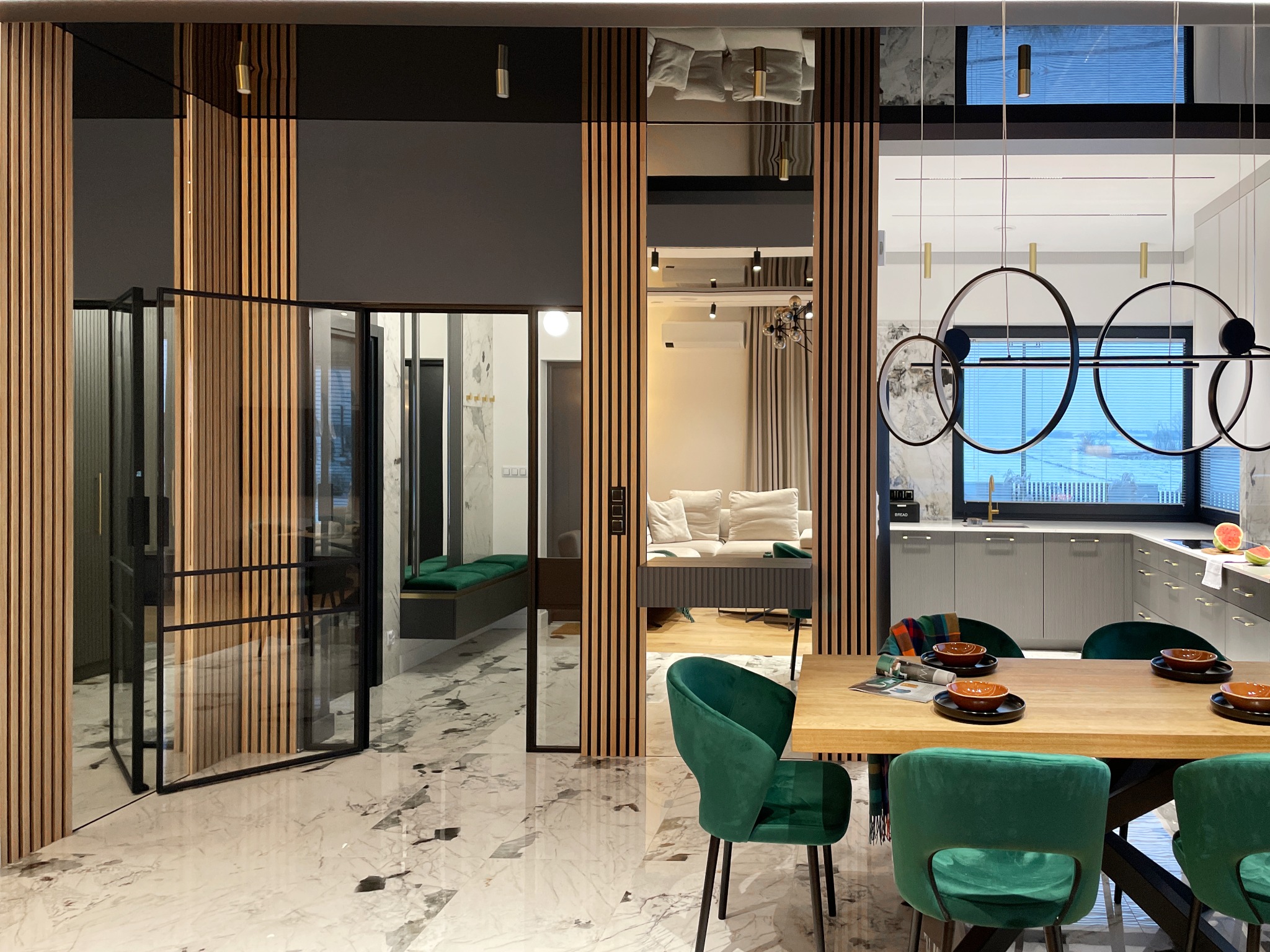 Nowoczesne wnętrze z marmurową podłogą, drewnianymi lamelami, szklanymi drzwiami i zielonymi krzesłami velvet przy drewnianym stole, widok na kuchnię z elementami złota.