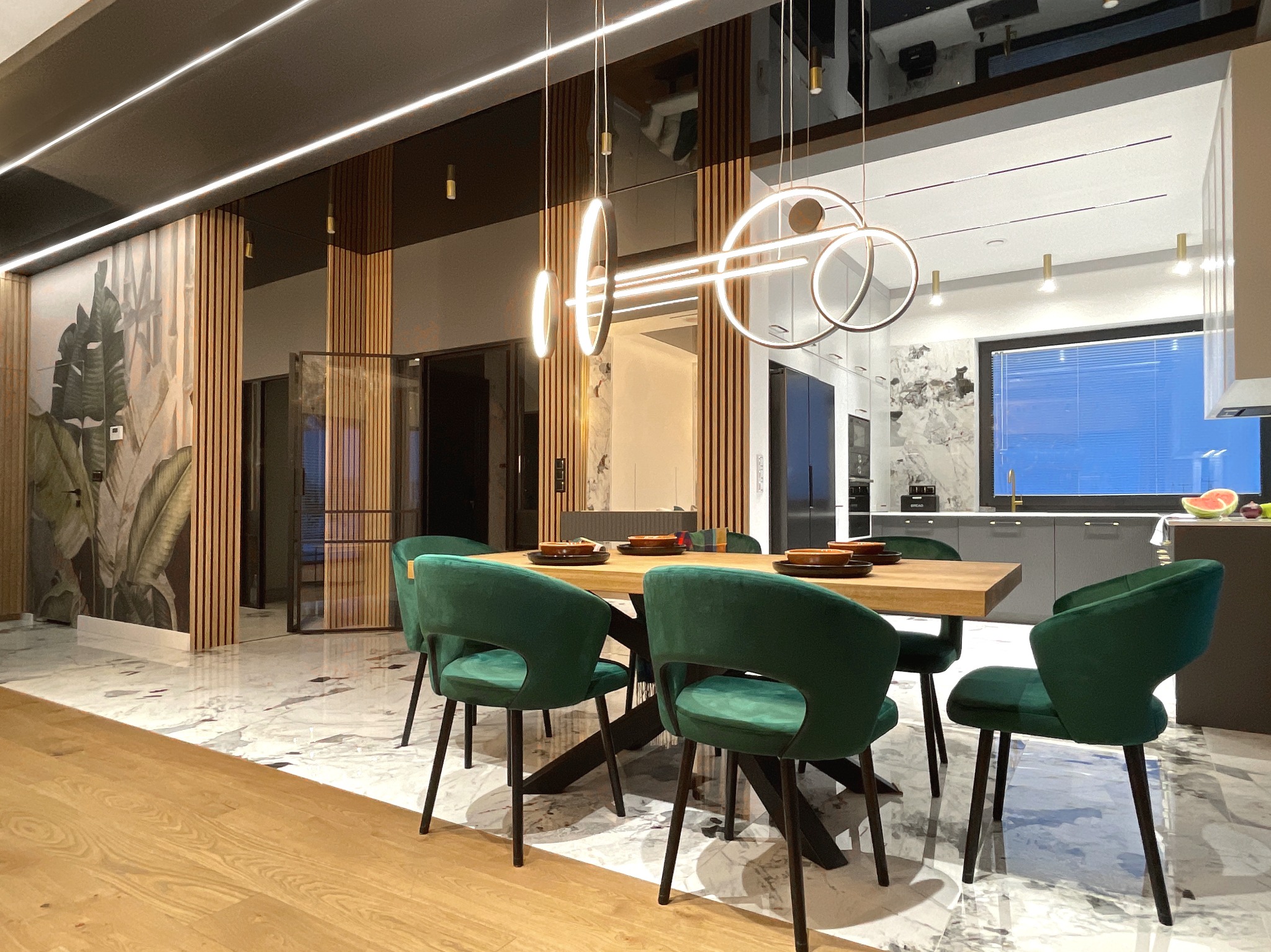 Nowoczesna jadalnia z drewnianym stołem, zielonymi krzesłami, marmurową podłogą i dekoracyjnym oświetleniem w postaci okrągłych lamp wiszących.
