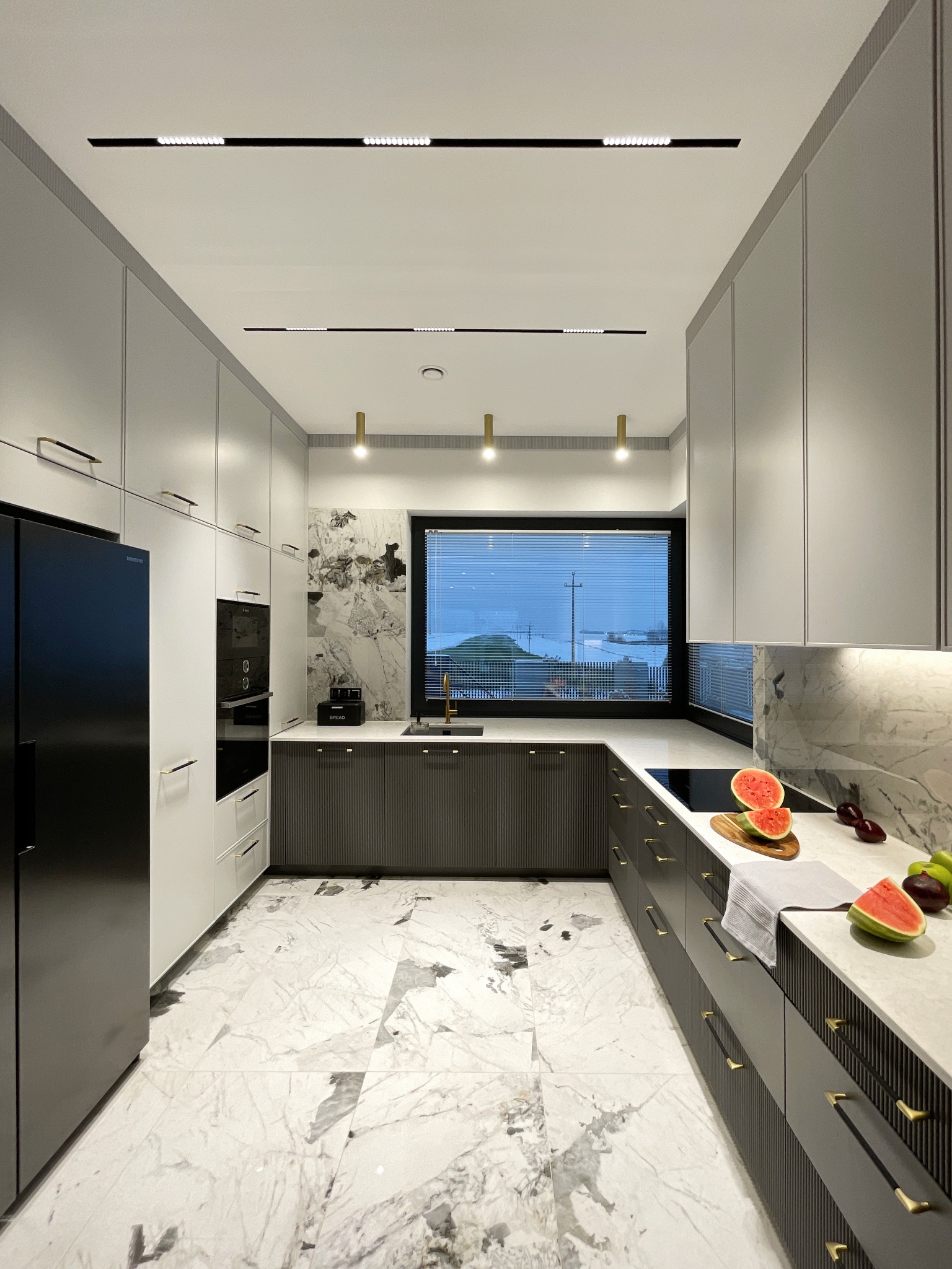 Nowoczesna kuchnia w odcieniach szarości i bieli z marmurową podłogą i blatem, widok na zimowy krajobraz za oknem, na blacie połówki arbuza i owoce.