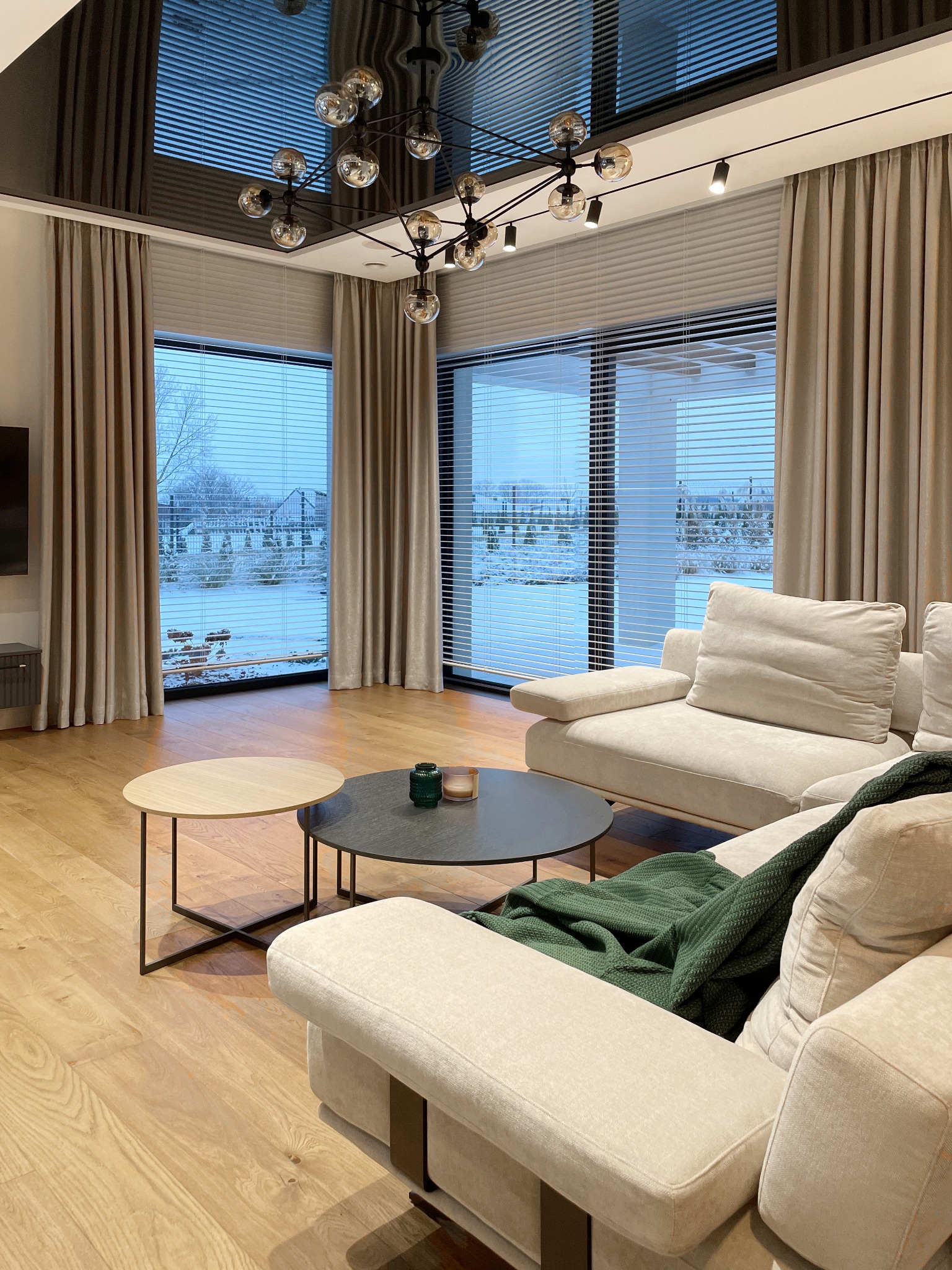 Jasny salon z dużymi oknami z widokiem na zimowy krajobraz, kremowa sofa z zielonym pledem, okrągłe stoliki kawowe, nowoczesna lampa wisząca z żarówkami w stylu Edison, zasłony w odcieniach beżu.