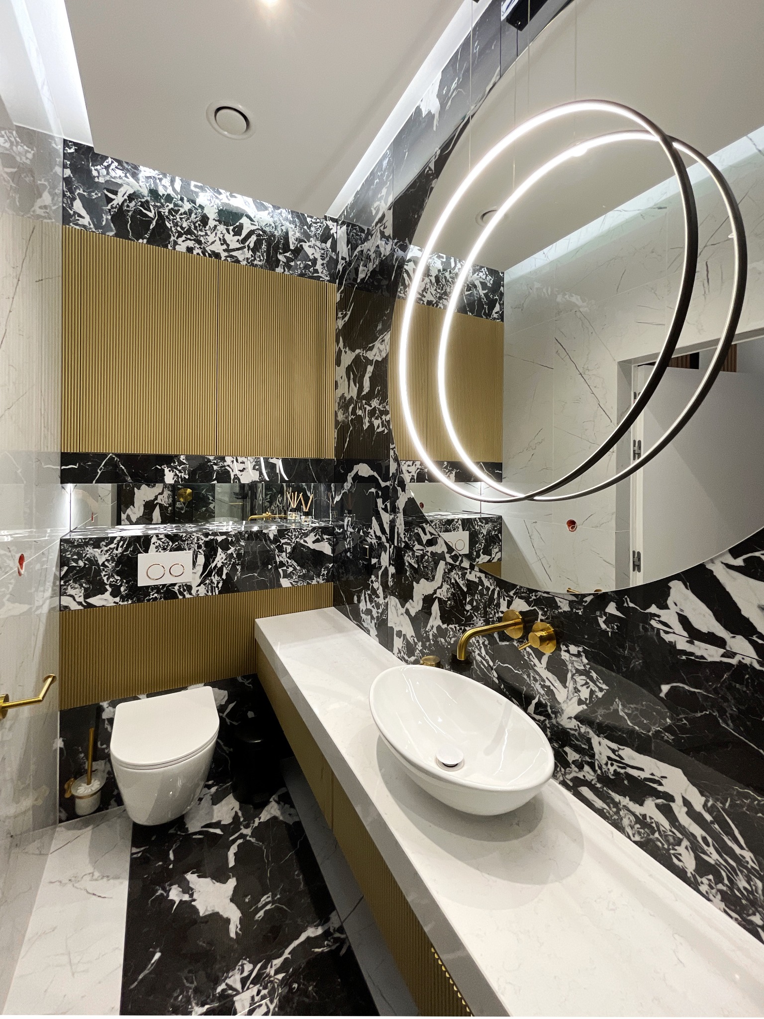 Nowoczesna łazienka z czarno-białymi marmurowymi płytkami, złotymi akcentami, wiszącą toaletą i umywalką nablatową na długim blacie, oświetlona okrągłym lustrem z LED.