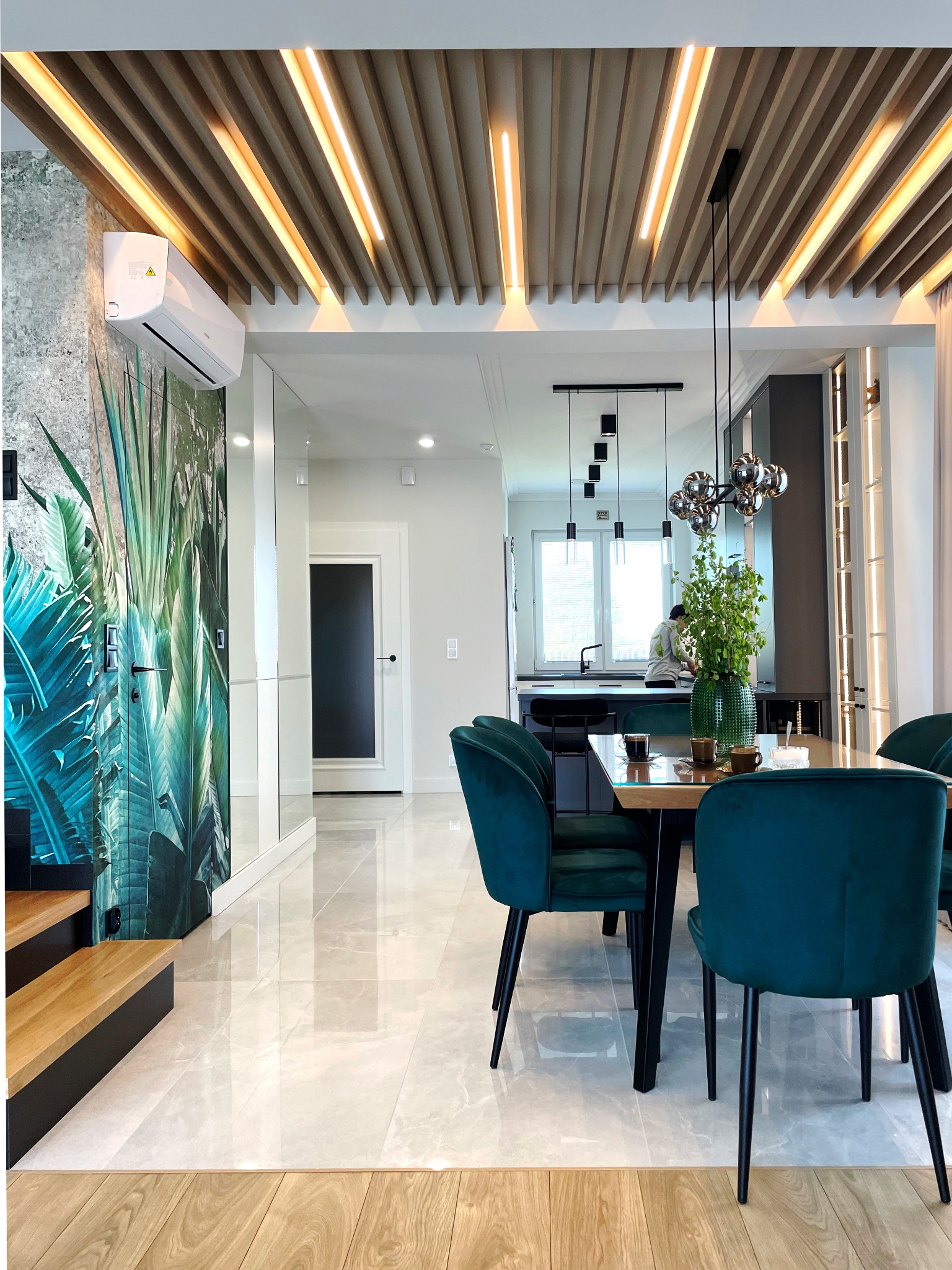 Jadalnia z drewnianym stołem, zielonymi krzesłami i dekoracyjnym sufitem z oświetleniem LED, widok na kuchnię w tle, ściana z motywem roślinnym i klimatyzator.