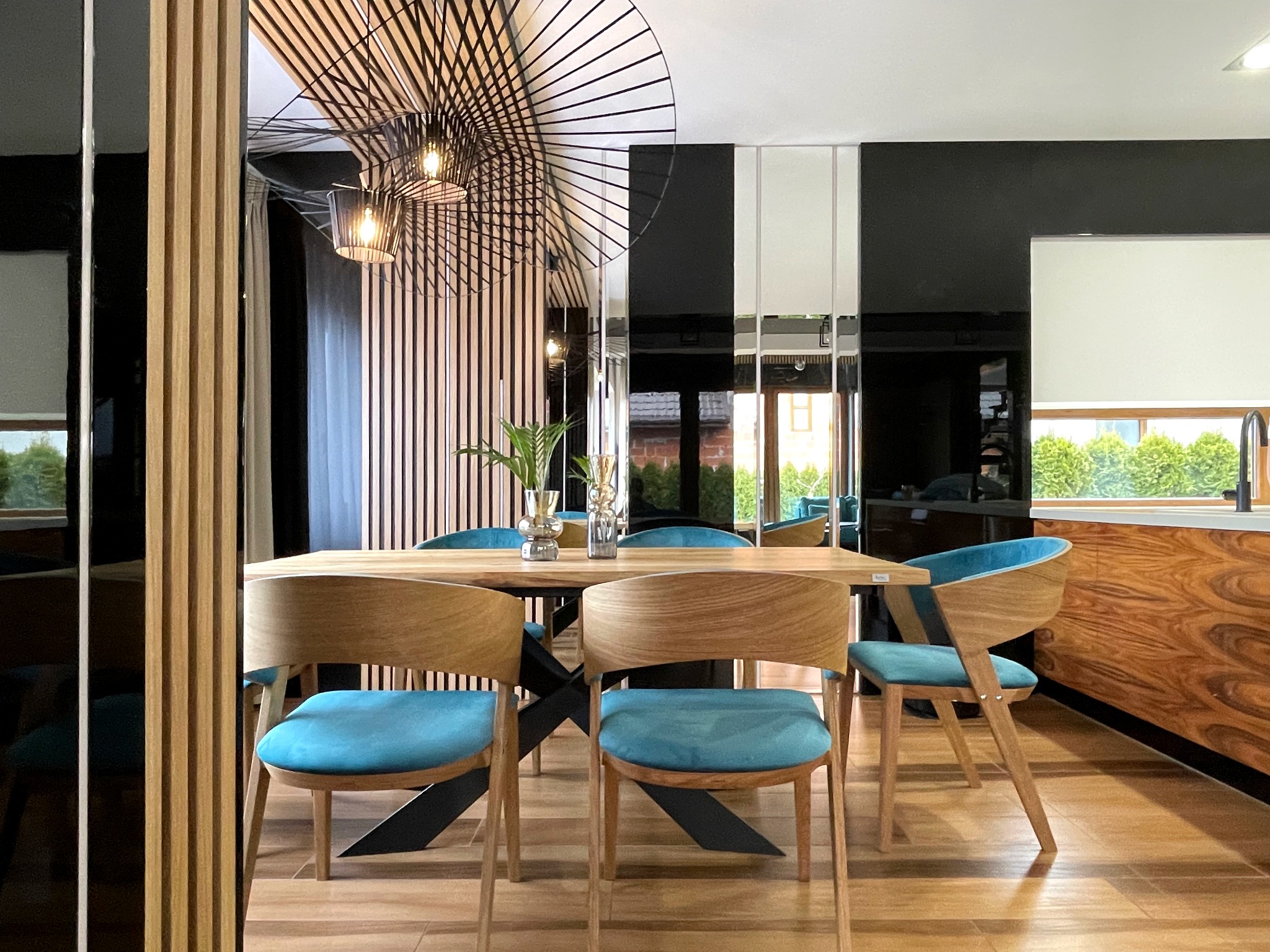 Nowoczesna jadalnia: drewniany stół z turkusowymi krzesłami, designerska lampa wisząca, drewniane lamele na ścianie i czarne, błyszczące panele.