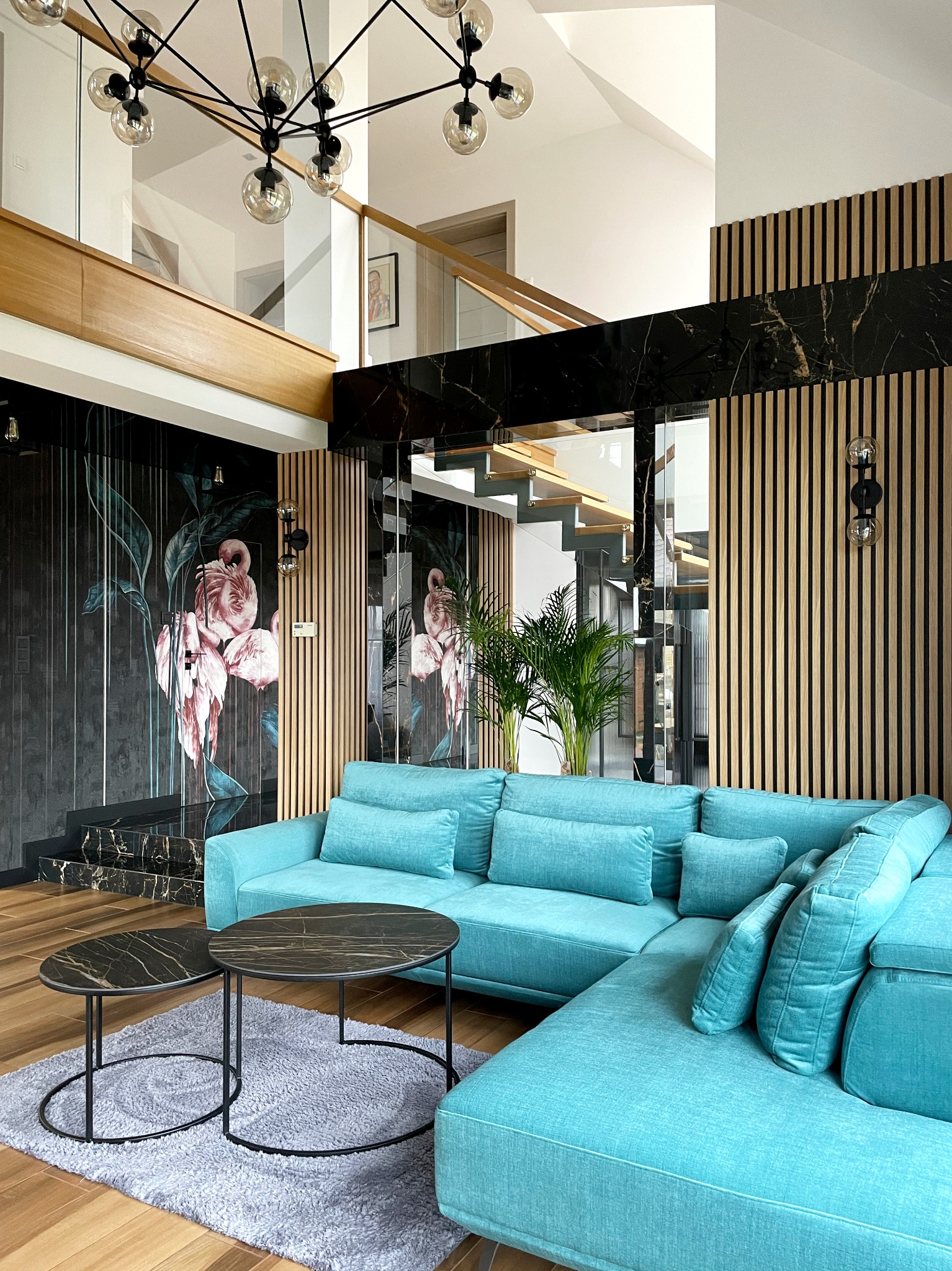 Turkusowy narożnik w salonie z akcentami czarnego marmuru i drewnianych lameli, ściana z motywem flamingów, nowoczesna lampa wisząca i schody z przeszkloną balustradą.