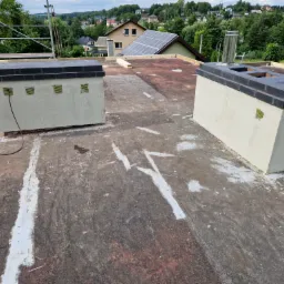 Płaski dach pokryty papą z dwoma kominami po renowacji, widoczne białe ślady po uszczelnianiu, w tle domy i zieleń.