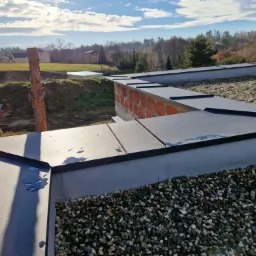 Nowoczesny dach z blachy na rąbek stojący, pokryty drobnym żwirem, z widocznym fragmentem muru i okolicznym krajobrazem Rogowa w tle, w słoneczny dzień.