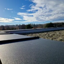 Nowoczesny dach z blachy trapezowej w kolorze grafitowym, widok z dołu na tle błękitnego nieba z chmurami, z widocznym fragmentem posypki kamiennej.