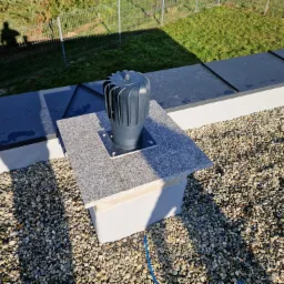 Szary odpowietrznik kominowy z turbiną na dachu pokrytym drobnymi kamieniami, osadzony na podstawie z płytek imitujących granit, widoczny cień osoby fotografującej.