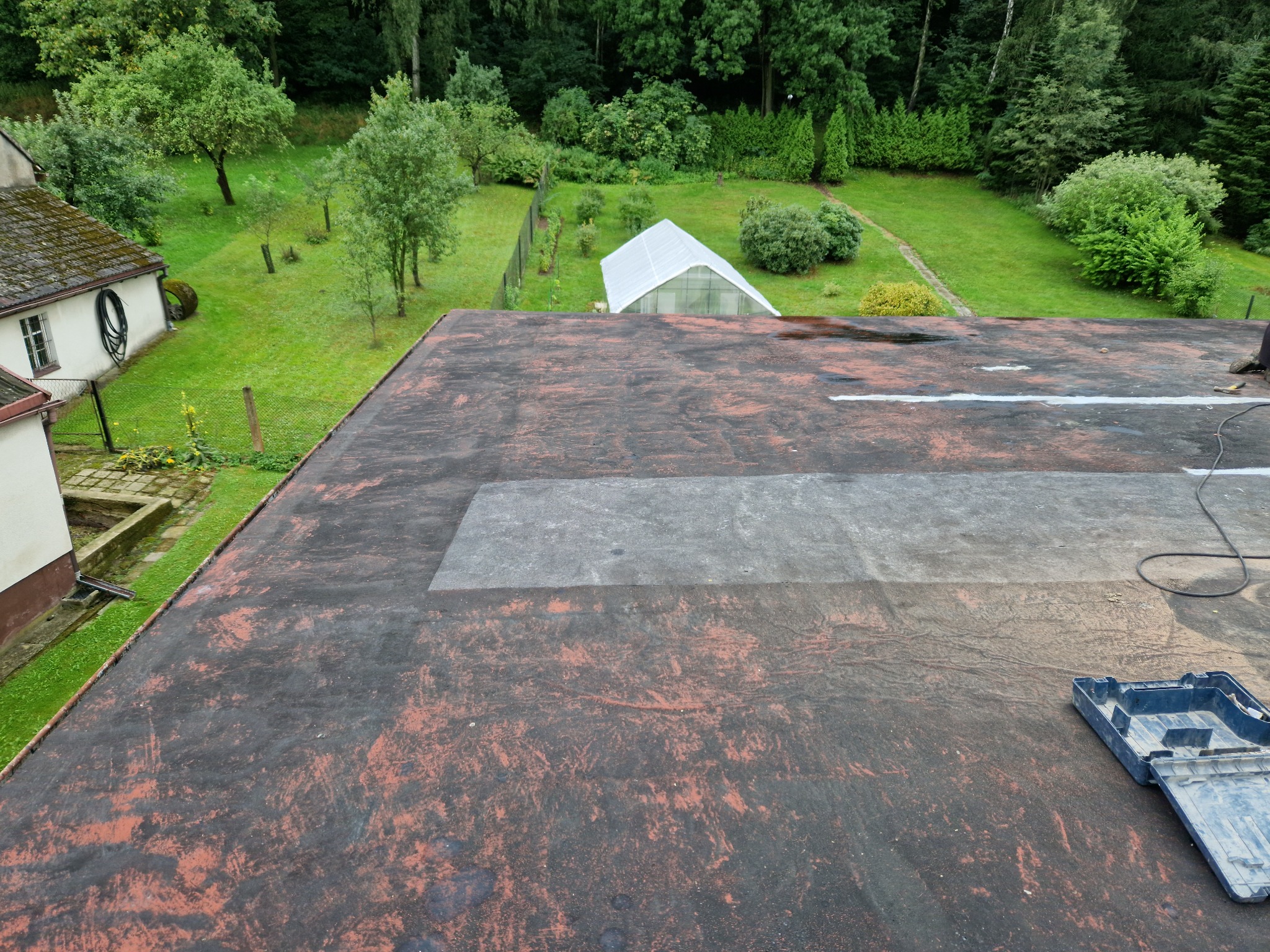 Płaski dach pokryty papą z widocznymi łatami, w tle ogród z tunelem foliowym i budynkiem gospodarczym.