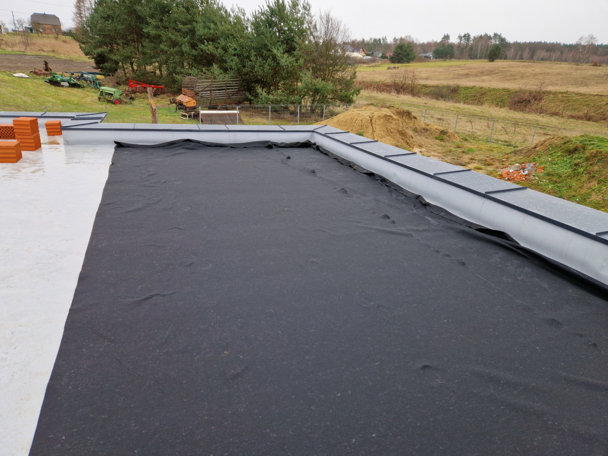 Widok z góry na fragment dachu pokrytego czarną membraną hydroizolacyjną, z widocznymi elementami obróbki blacharskiej na krawędzi, w tle krajobraz wiejski z drzewami i polami.
