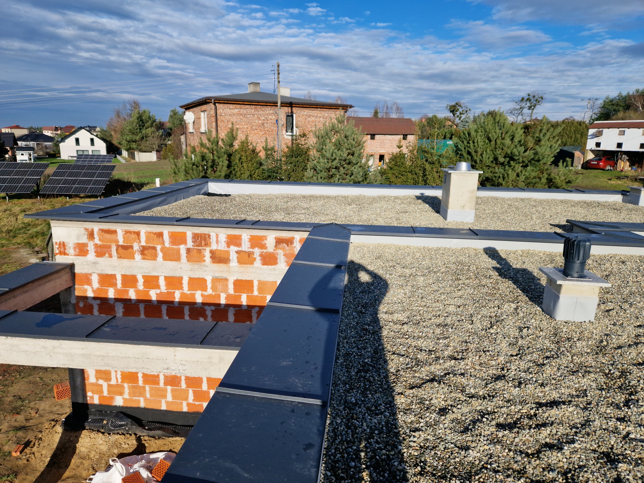 Płaski dach pokryty drobnym żwirem, z widocznymi elementami obróbki blacharskiej i kominem wentylacyjnym, w tle zabudowa jednorodzinna i panele solarne.