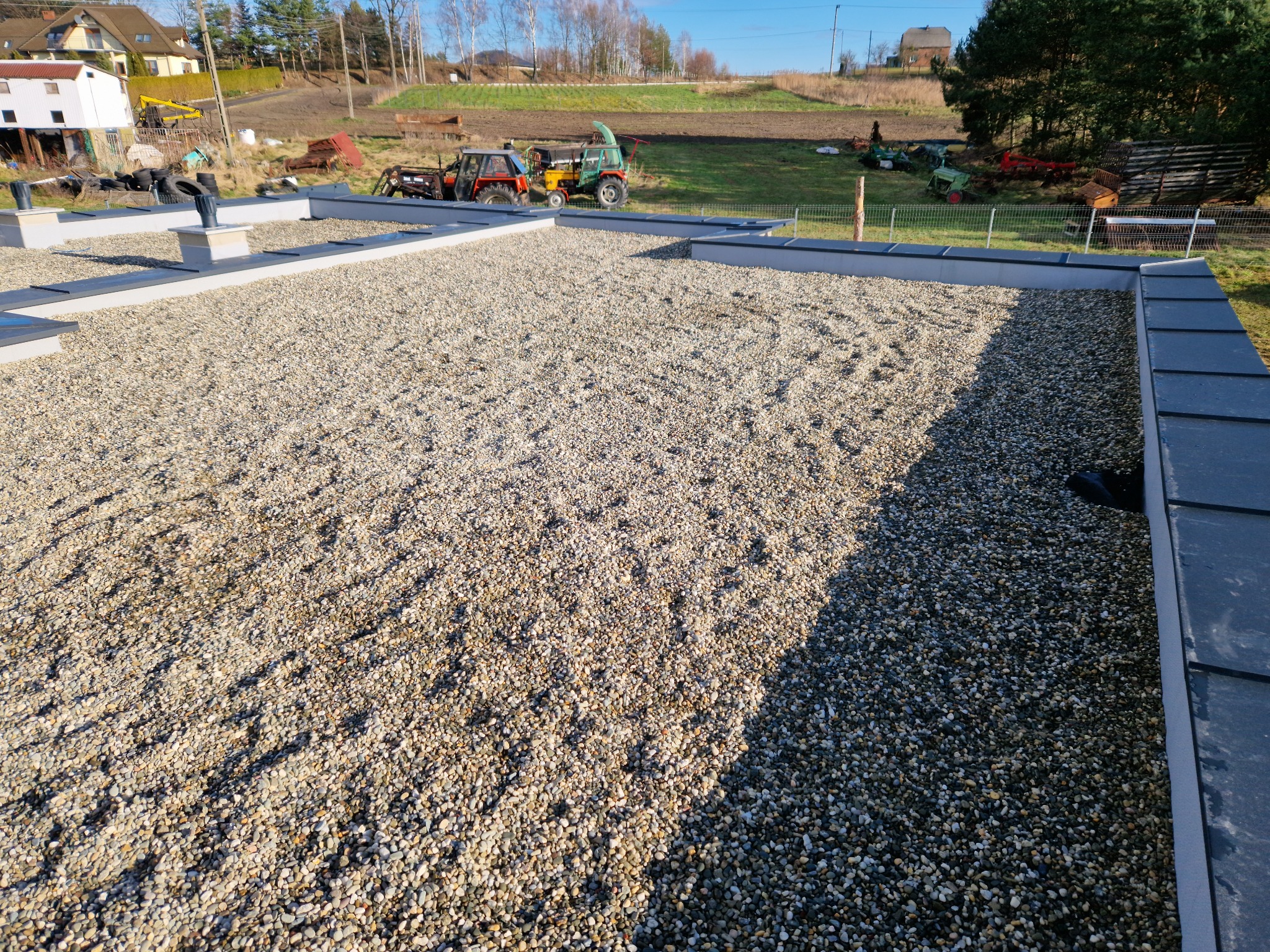 Nowoczesny dach płaski pokryty drobnym, jasnym żwirem, z widocznymi elementami wentylacji i obróbkami blacharskimi, w tle zabudowania gospodarcze i pole uprawne.