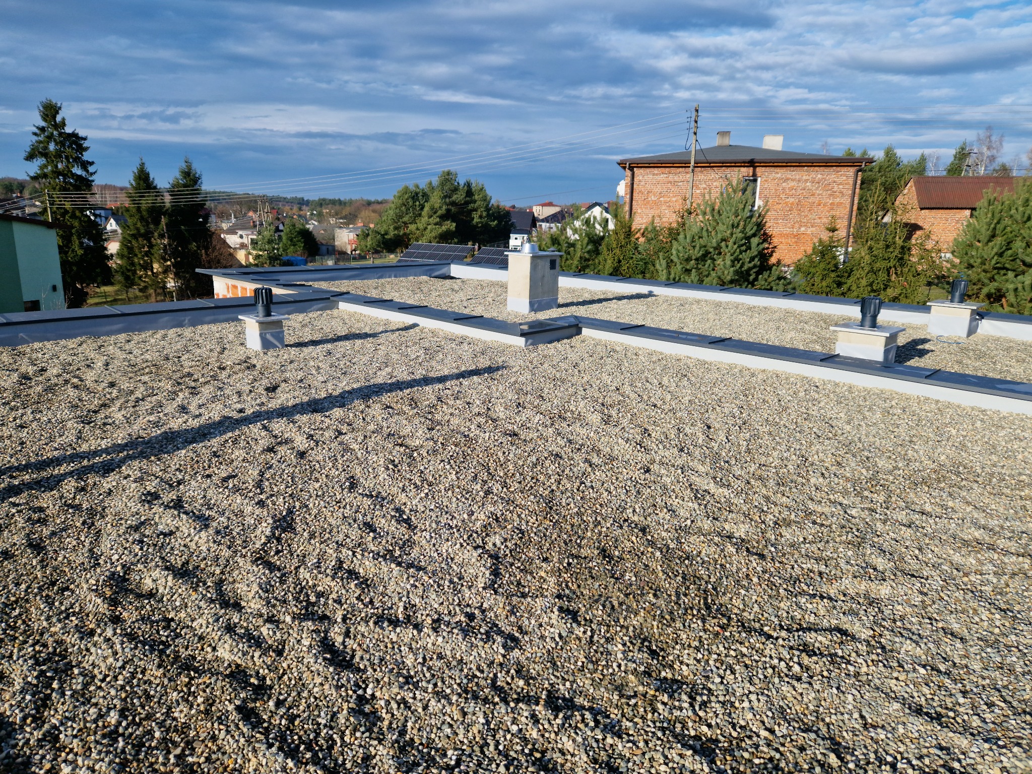 Płaski dach pokryty drobnym żwirem, z widocznymi elementami wentylacyjnymi i obróbkami blacharskimi, w tle zabudowa jednorodzinna i błękitne niebo z chmurami.