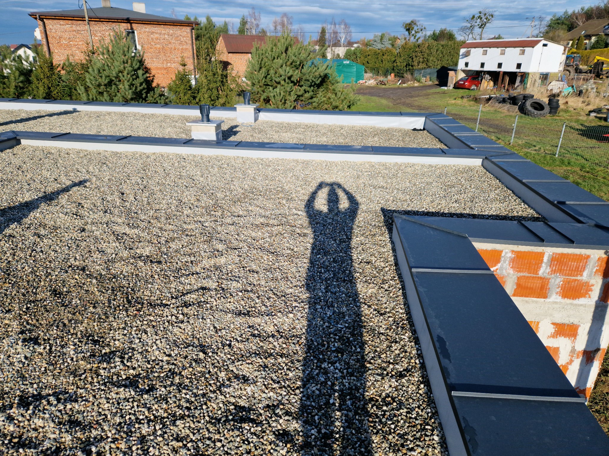 Nowoczesny dach płaski pokryty drobnym żwirem, z widoczną obróbką blacharską w kolorze grafitowym i cieniem osoby wykonującej zdjęcie, w tle zabudowa jednorodzinna i zieleń.