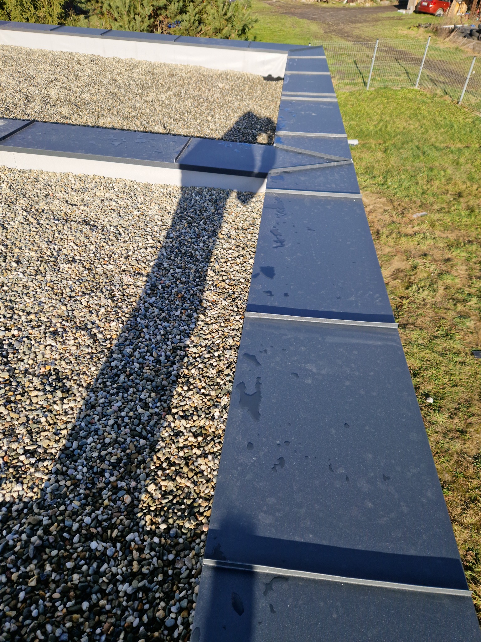 Widok z góry na fragment dachu pokrytego blachą w kolorze grafitowym, sąsiadującego z fragmentem dachu pokrytego drobnym żwirem. Widoczne łączenia blachy oraz cień rzucany na żwir.