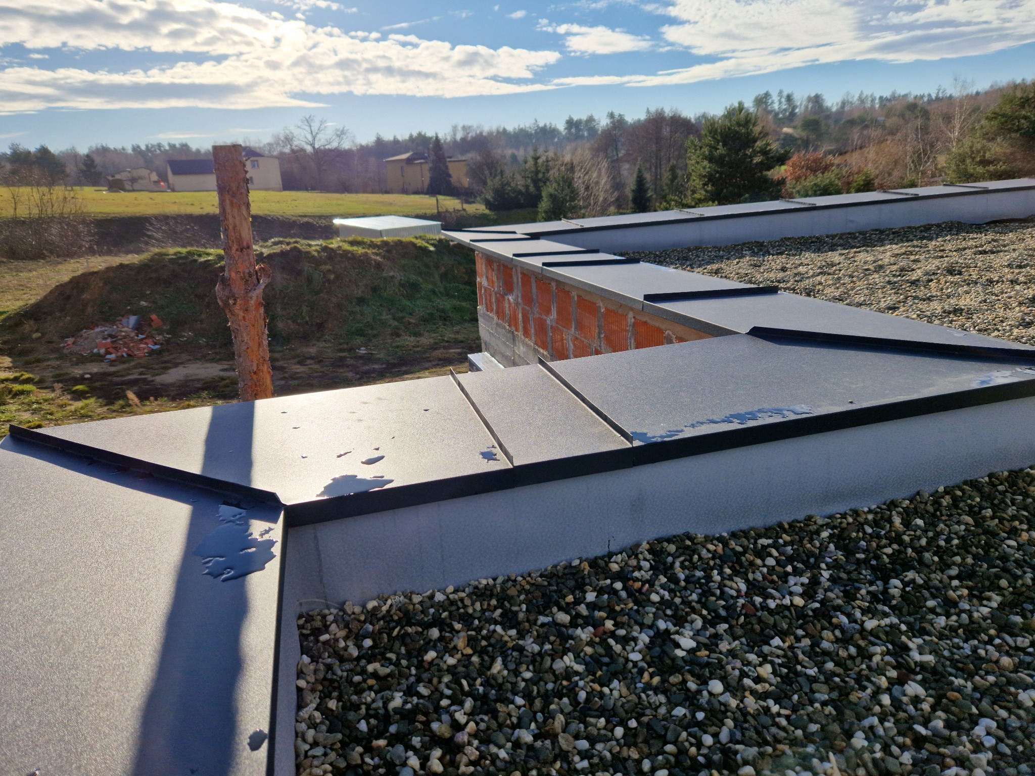 Nowoczesny dach z blachy na rąbek stojący, pokryty drobnym żwirem, z widocznym fragmentem muru i okolicznym krajobrazem Rogowa w tle, w słoneczny dzień.