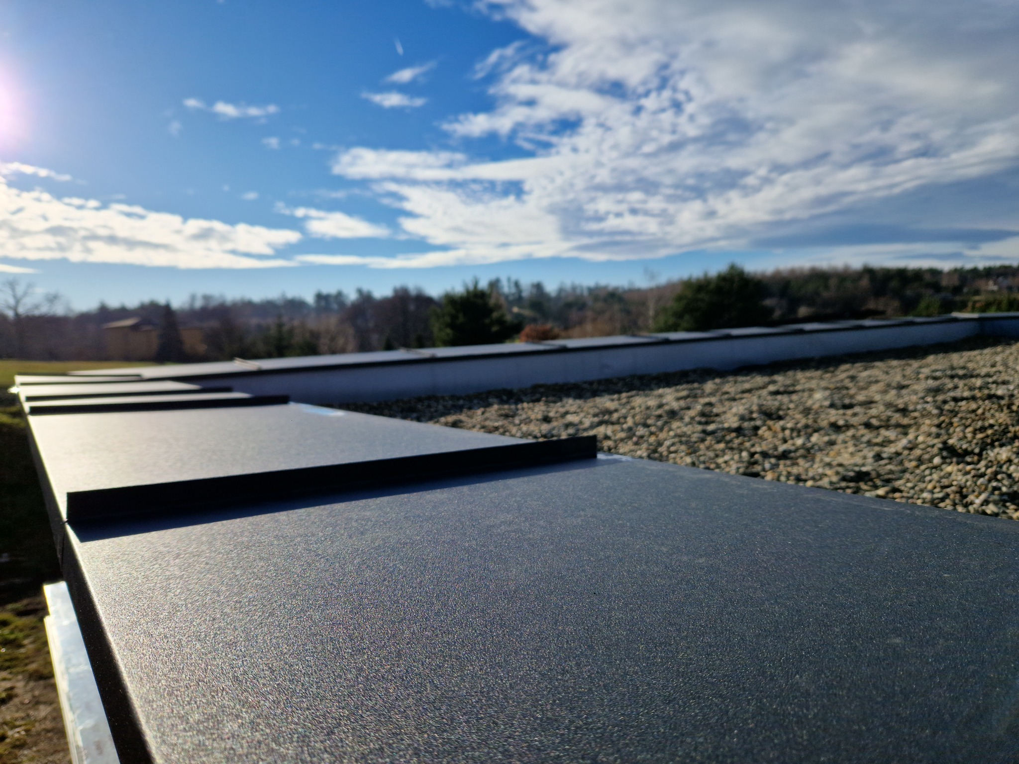 Nowoczesny dach z blachy trapezowej w kolorze grafitowym, widok z dołu na tle błękitnego nieba z chmurami, z widocznym fragmentem posypki kamiennej.