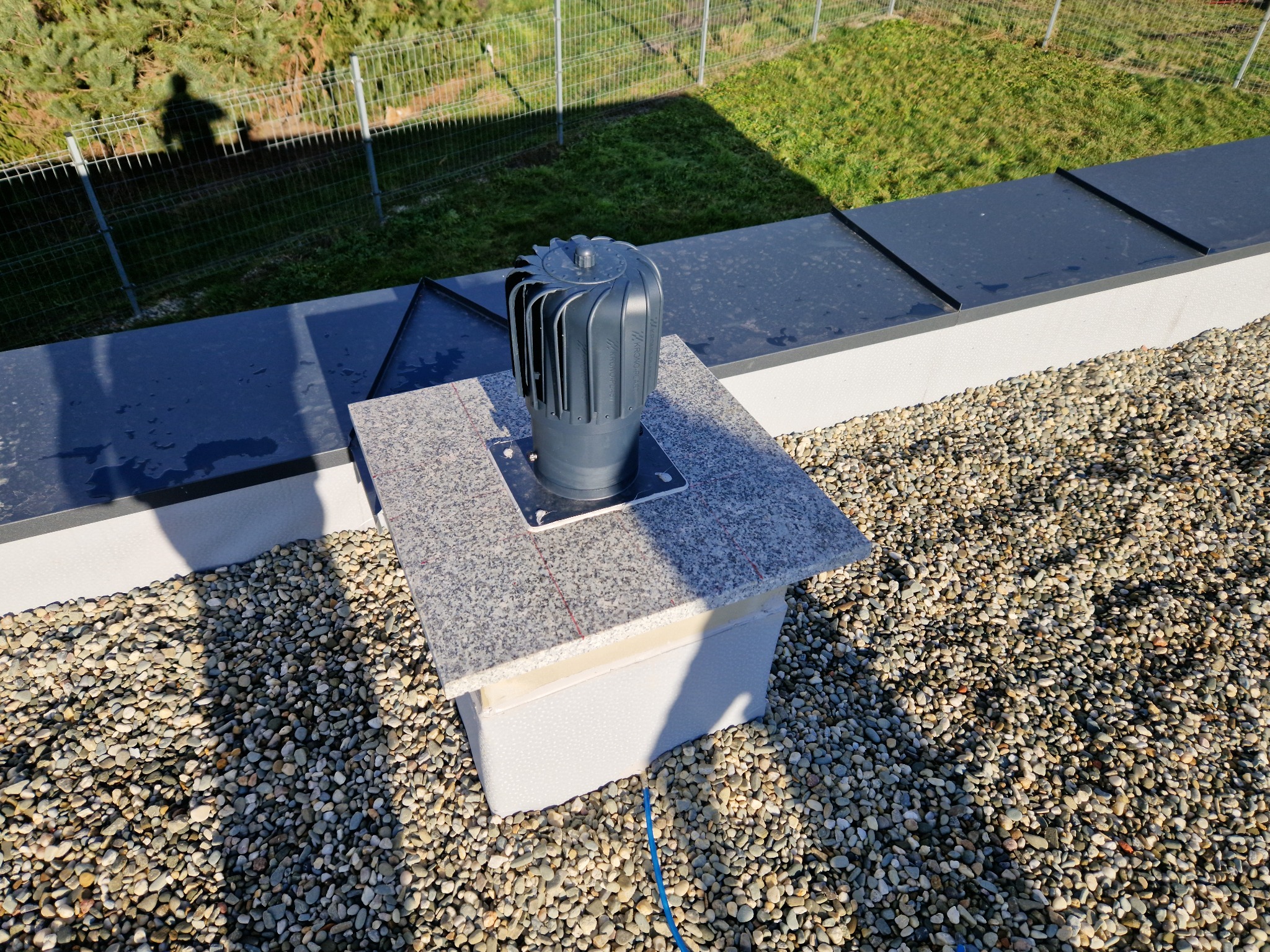 Szary odpowietrznik kominowy z turbiną na dachu pokrytym drobnymi kamieniami, osadzony na podstawie z płytek imitujących granit, widoczny cień osoby fotografującej.